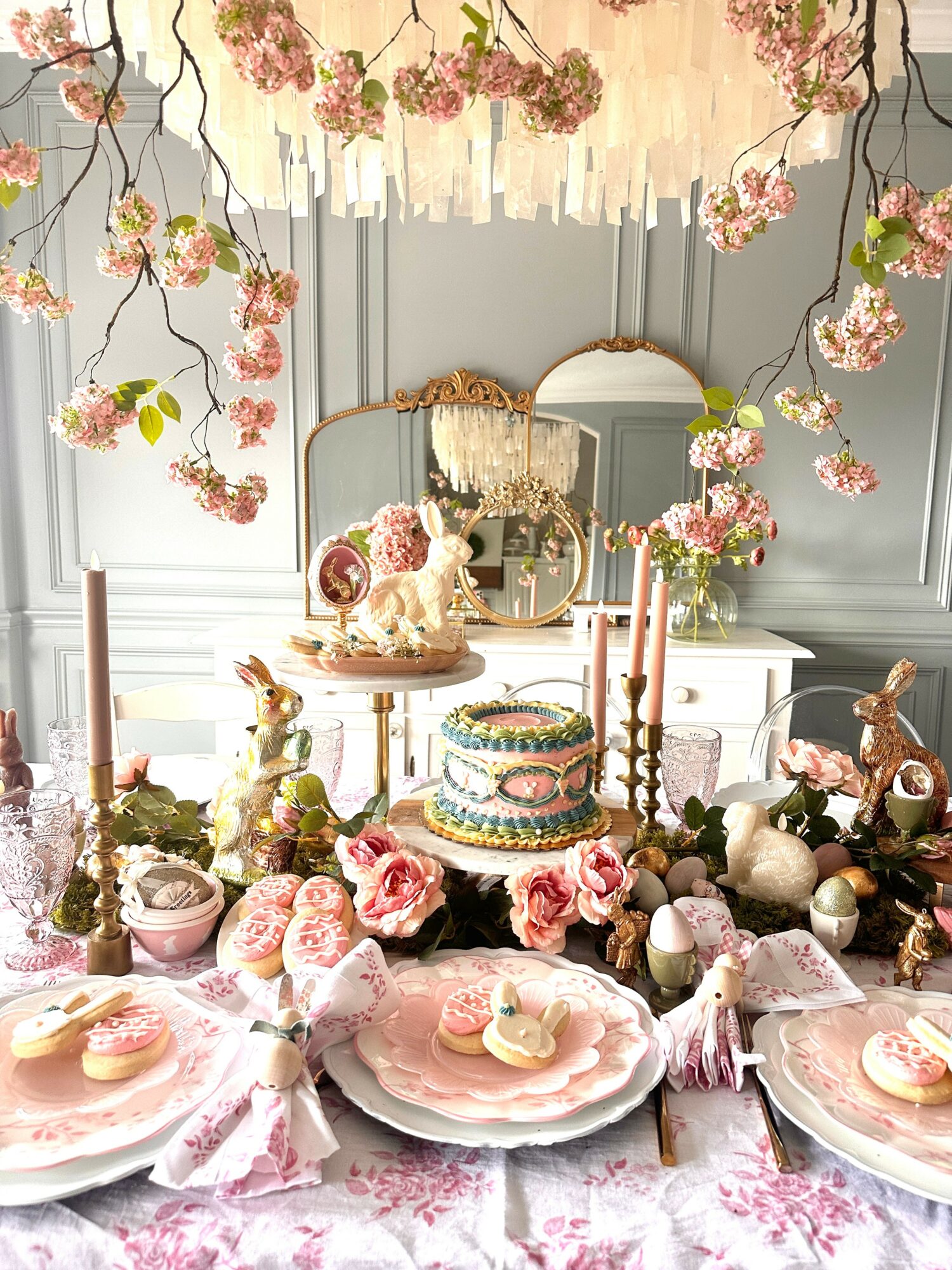 Decorative table with pink and white desserts, flowers, and a cake, set in a room with floral wall art and a mirror.