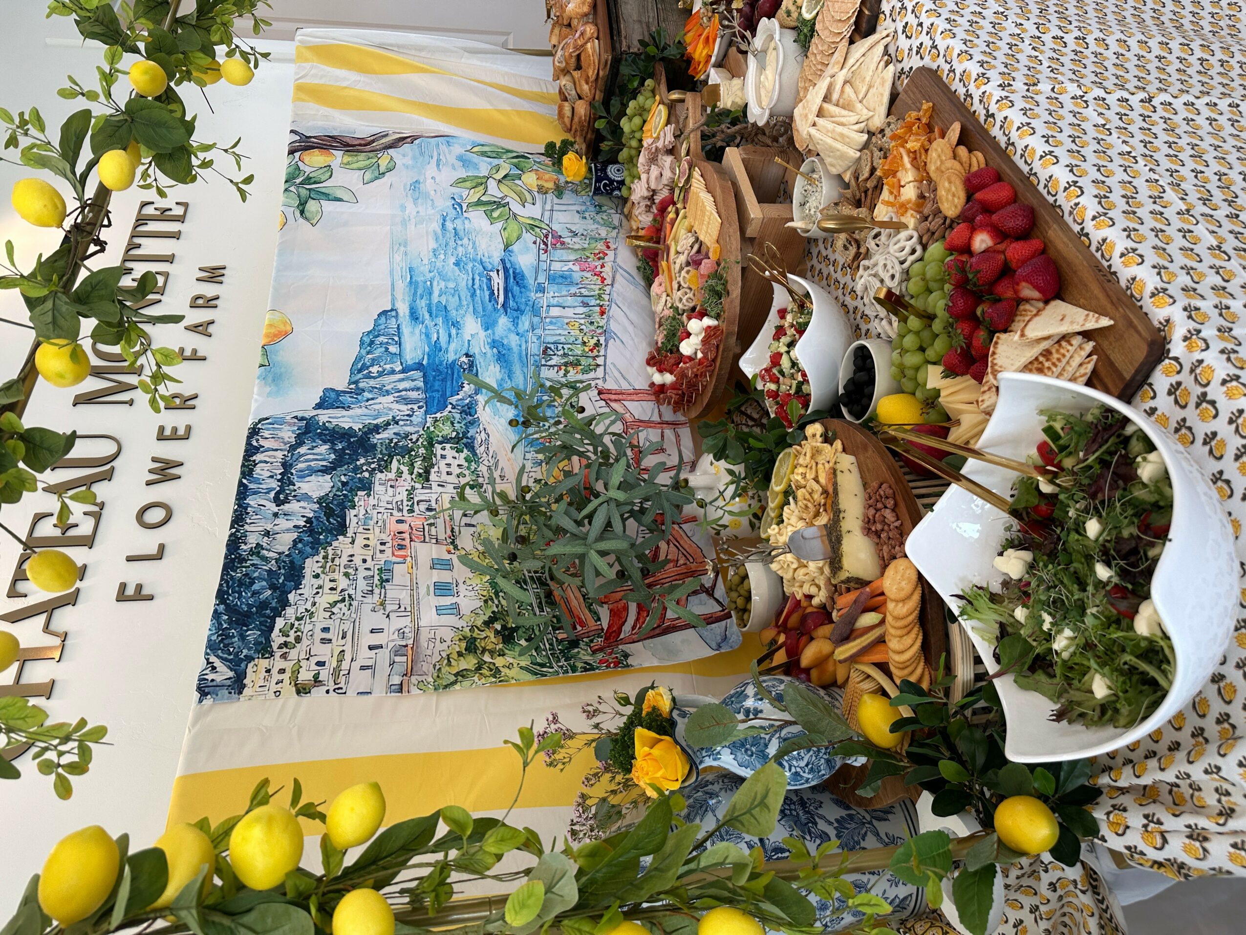 Display of flowers, fruits, and decorative items on a table with a cityscape backdrop, featuring yellow and green colors.
