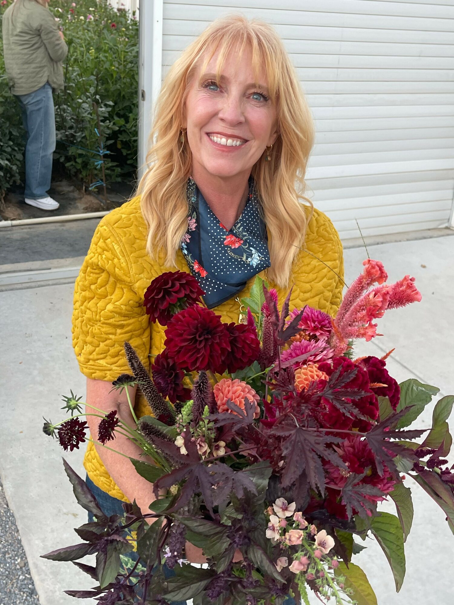 Woman with blonde hair smiling, holding a large colorful bouquet of flowers, wearing a yellow jacket and blue scarf.