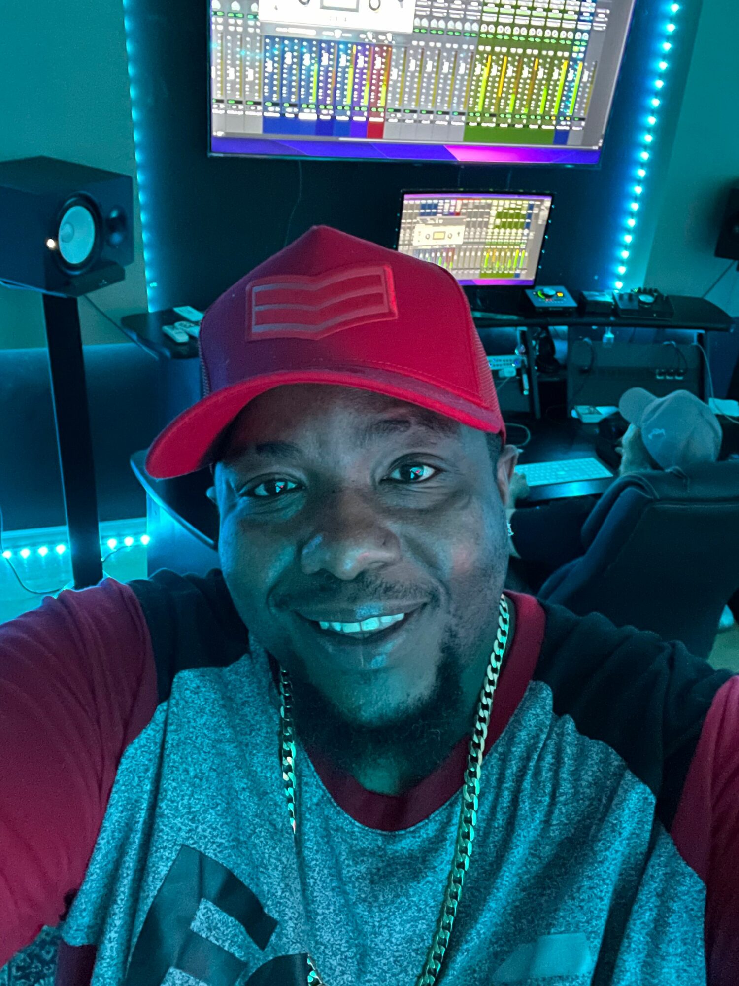 Person wearing a red cap and gray shirt smiling in a gaming or control room with screens and equipment.