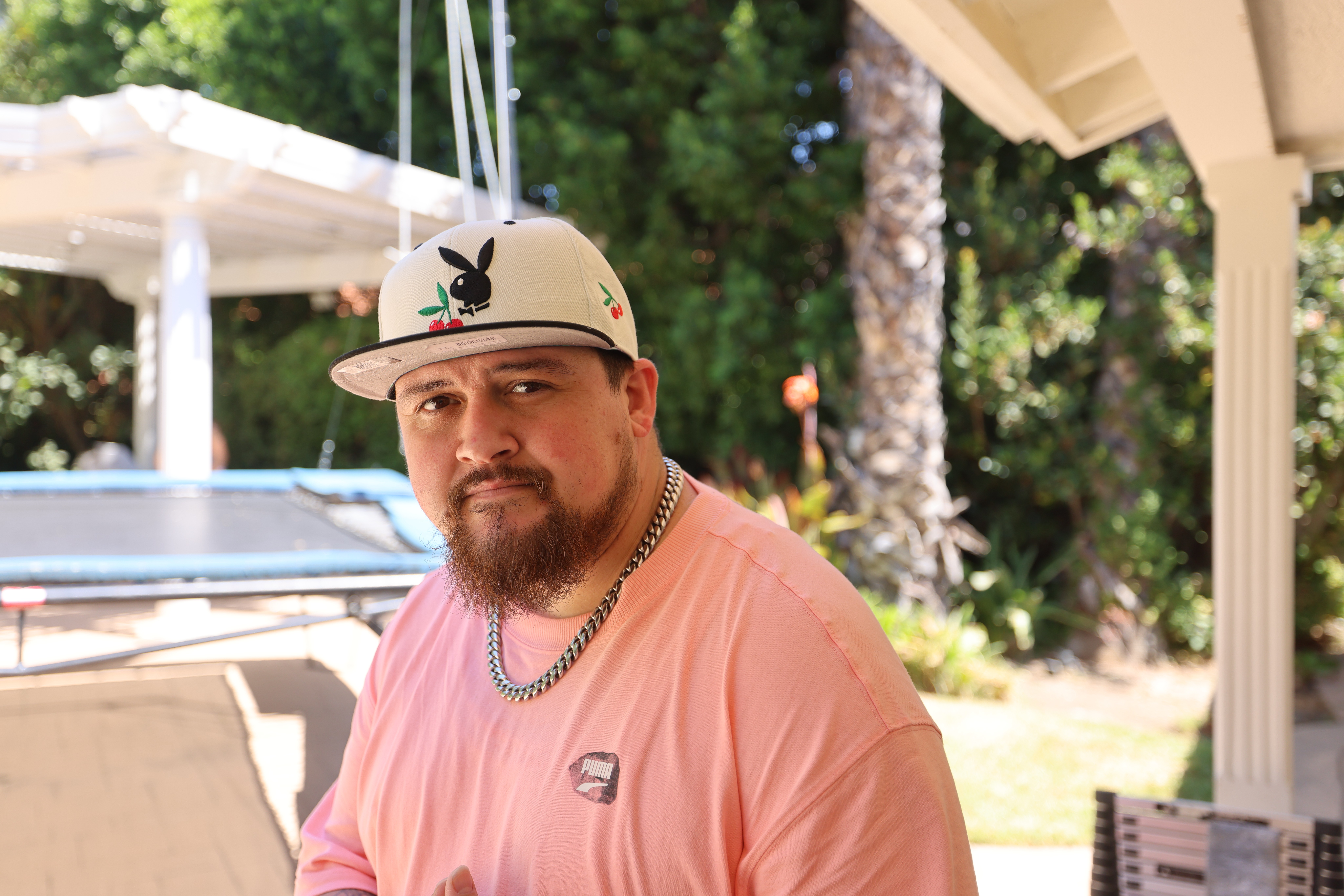 Man with a beard wearing a white cap with a black logo, pink shirt, and chain necklace outdoors near trees and a boat.