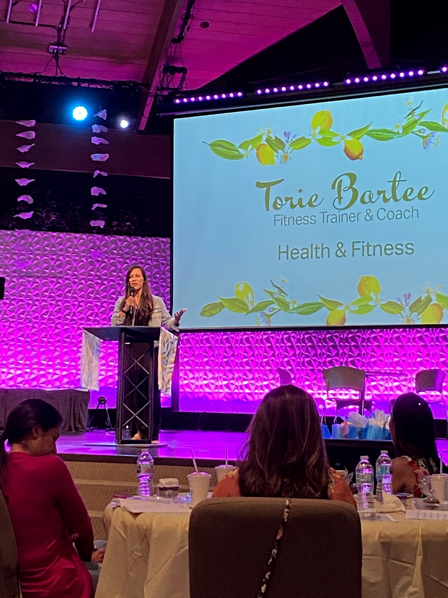 Woman standing at podium on stage with large screen displaying text and decorative leaves, audience seated at tables, purple lighting.