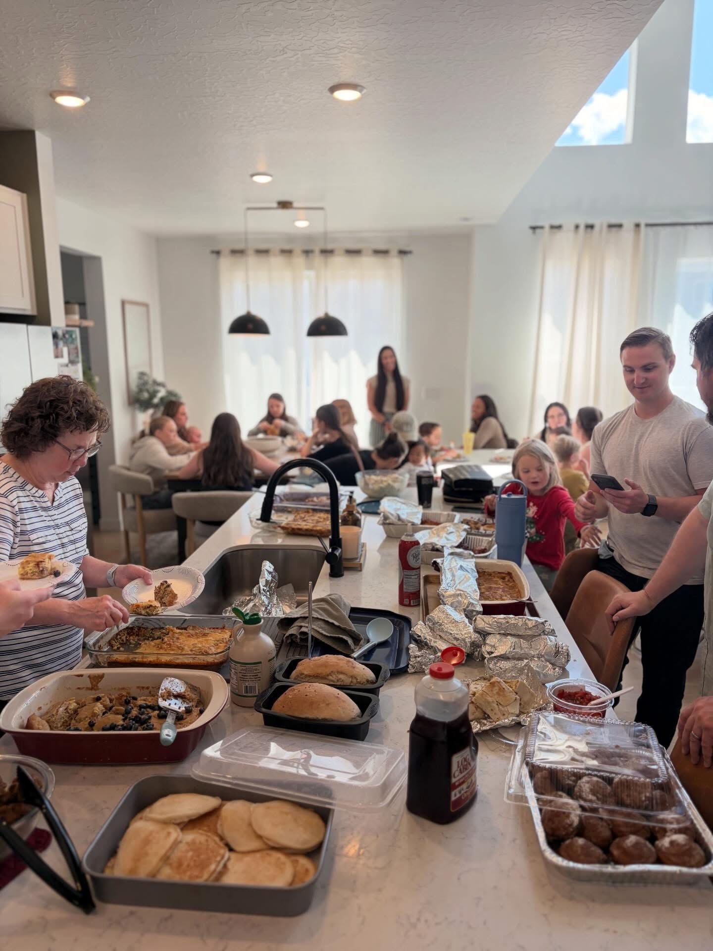 People gather in a bright kitchen with a breakfast bar, serving food and socializing, with a group of children and adults present.