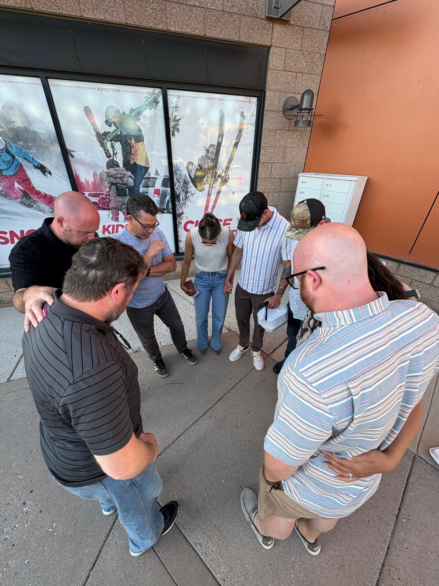 Group of people standing outside near a glass window, some looking at a display, others talking, with a person in a striped shirt in the foreground.