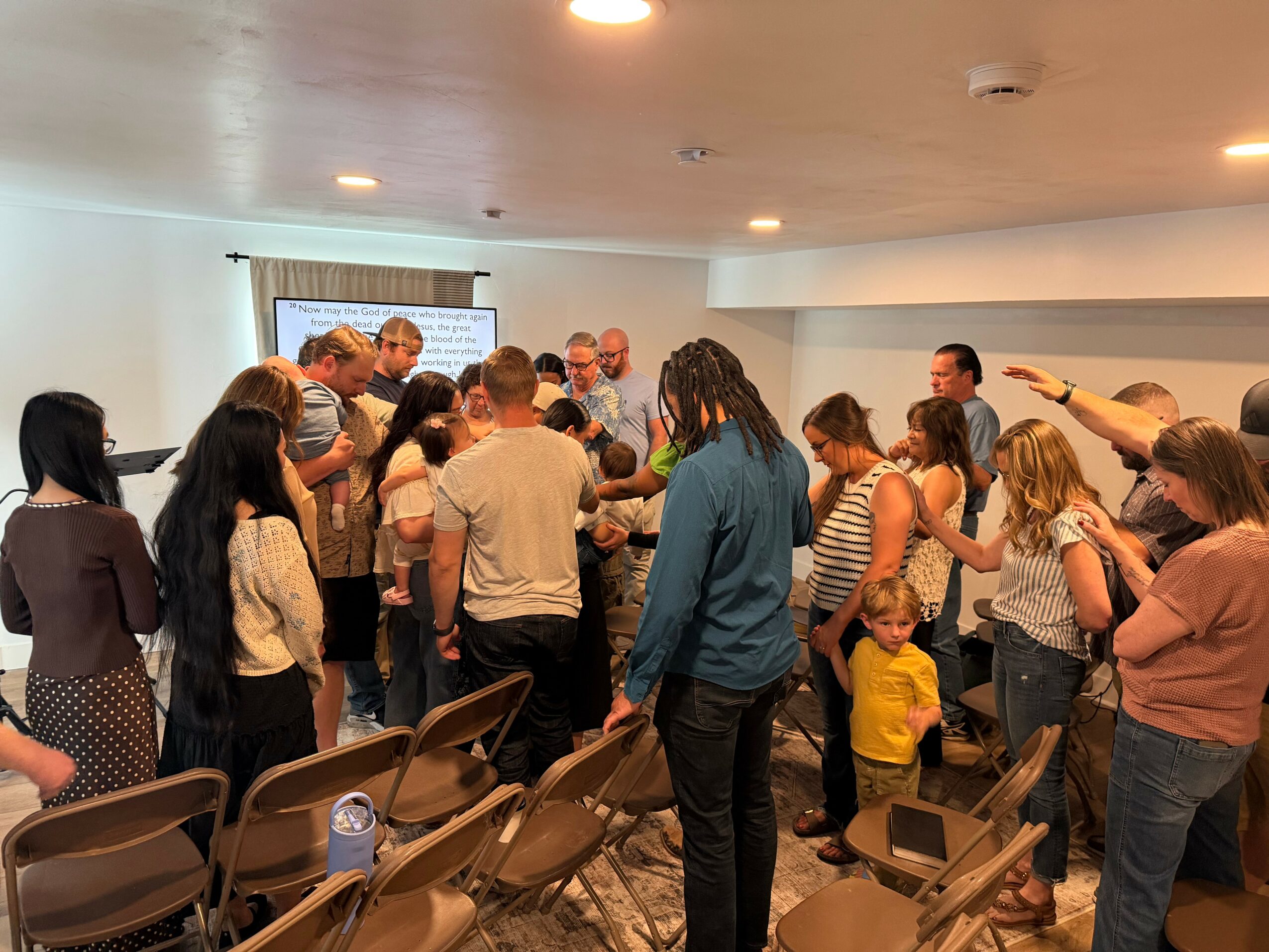 Group of people gathered in a room, some standing and some sitting, with chairs and a window in the background.