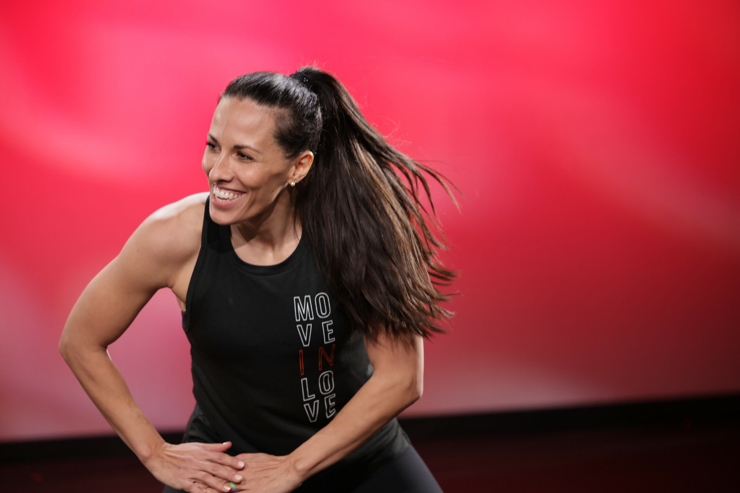 Woman with long hair smiling, wearing a black tank top, standing against a red background.