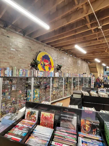 Bookshelves filled with comic books and graphic novels in a store, brick wall with a colorful logo, ceiling with wooden beams.