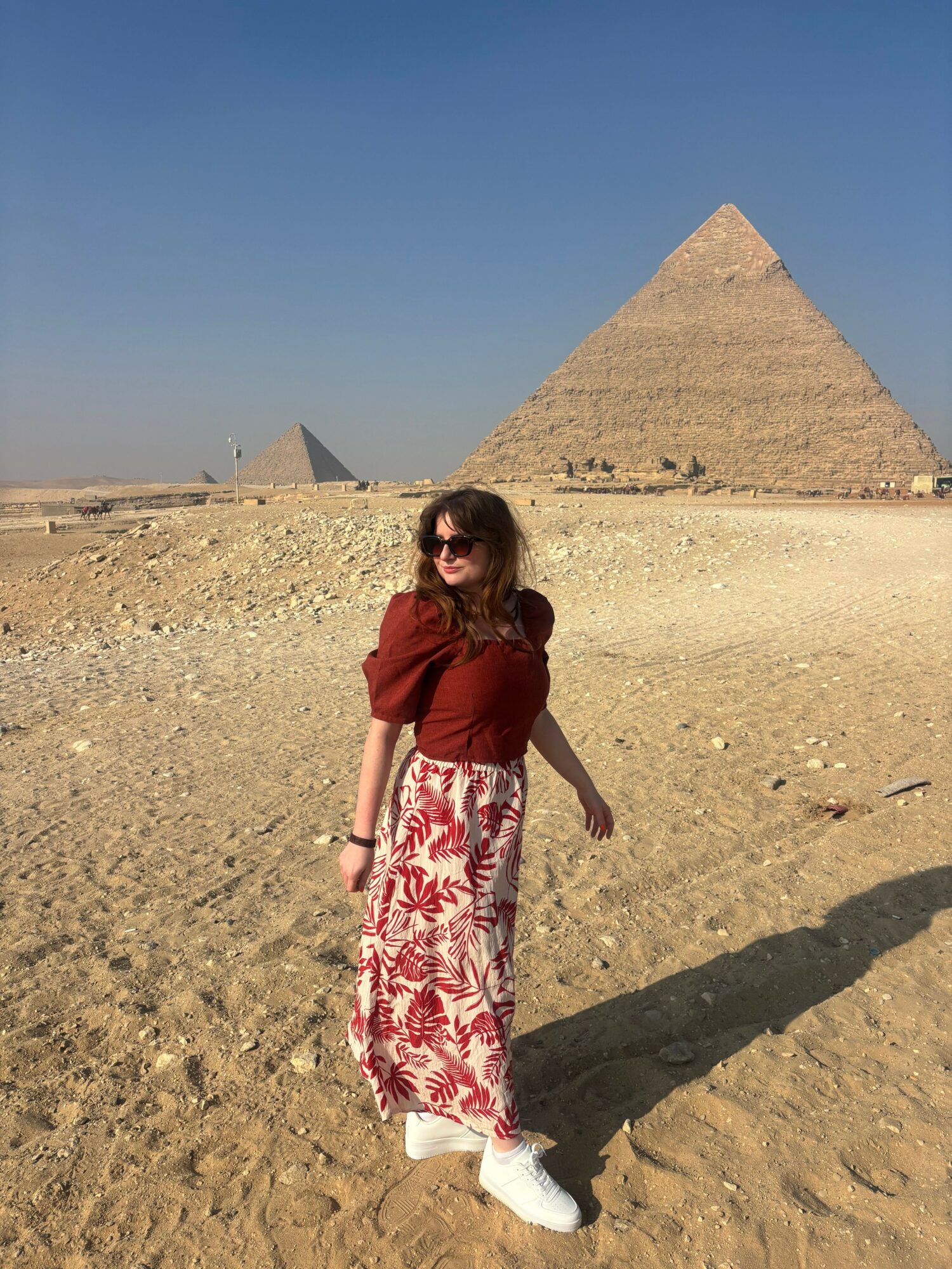 Woman in red top and patterned skirt walking in desert with pyramid in background.