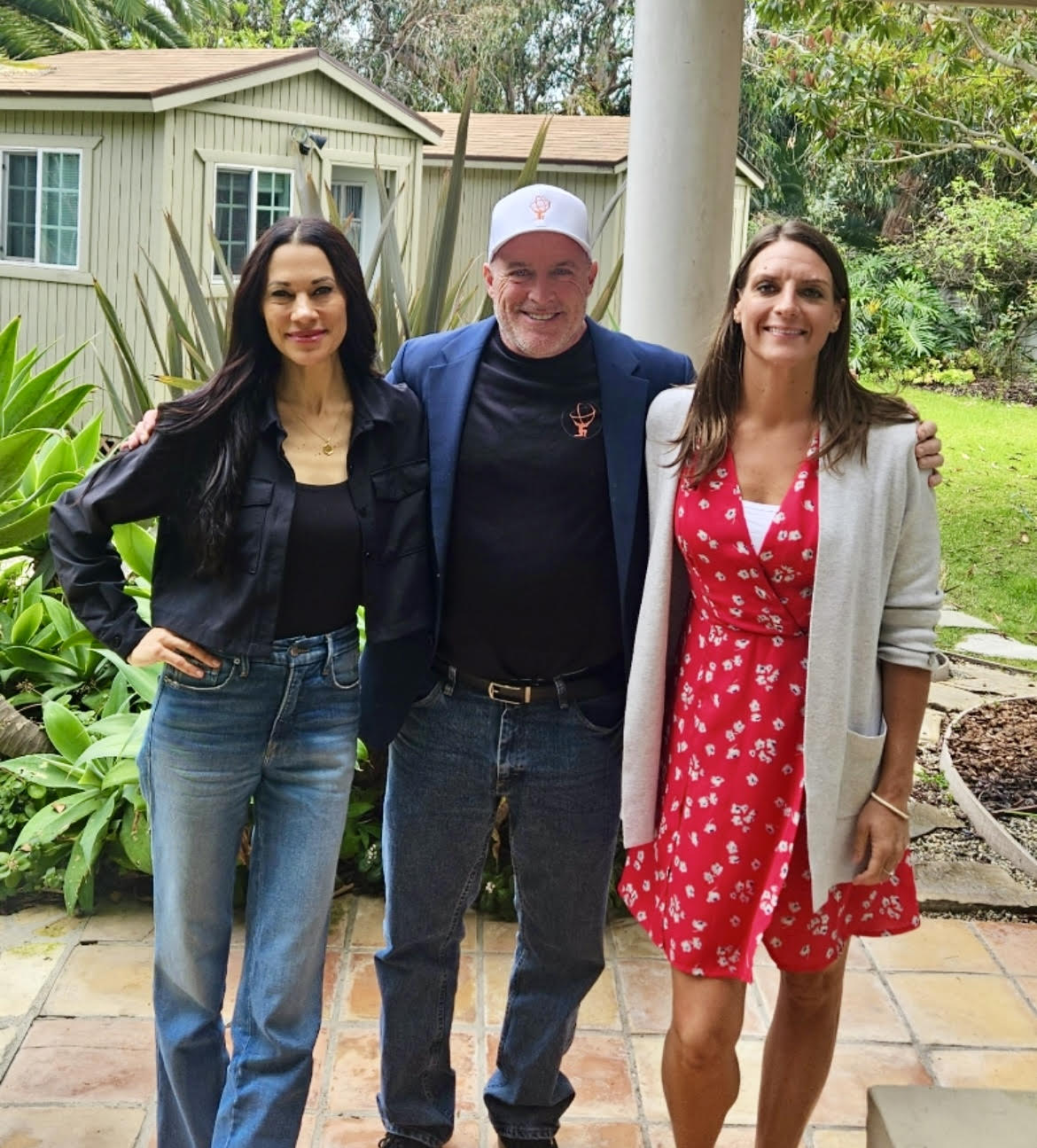 Three people standing outdoors on a patio, smiling, with greenery and a house in the background.