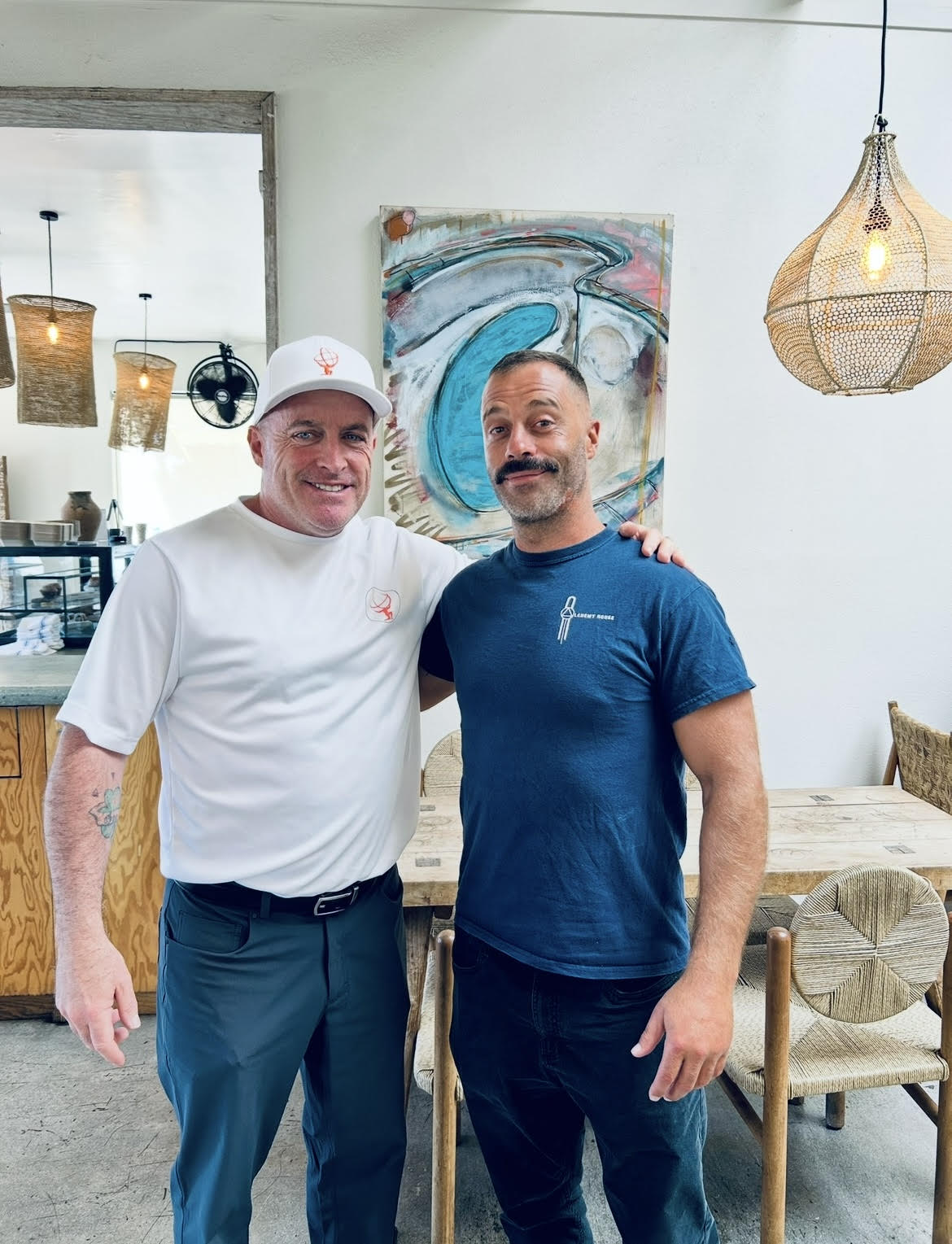 Two men stand together in a cafe, smiling, with artwork and hanging lights in the background.