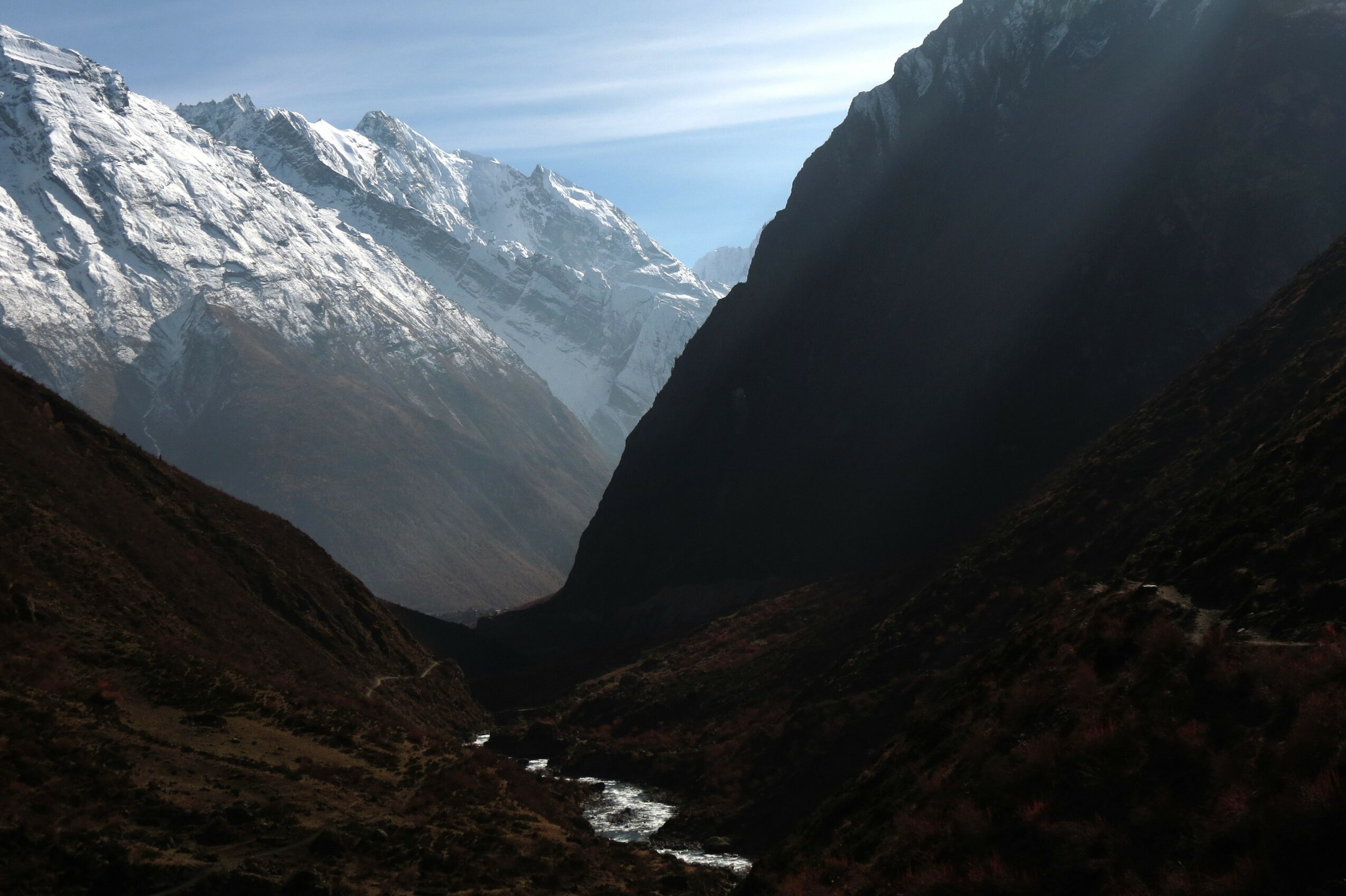 Mountain valley with snow-capped peaks, steep dark slopes, and a small river flowing through the valley floor.