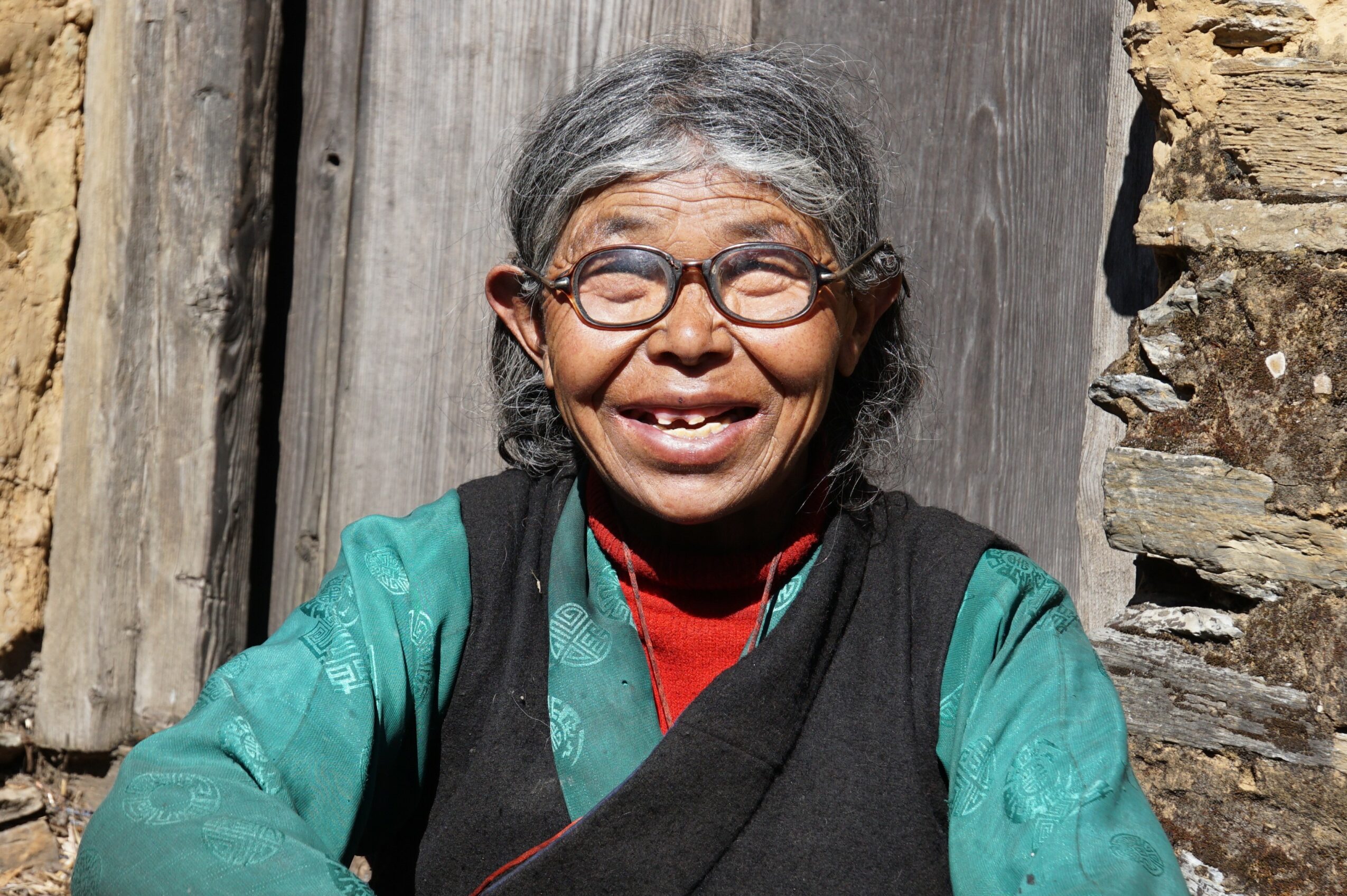 Smiling elderly woman with glasses, gray hair, wearing a teal jacket and red shirt, outdoors near wooden and stone structures.