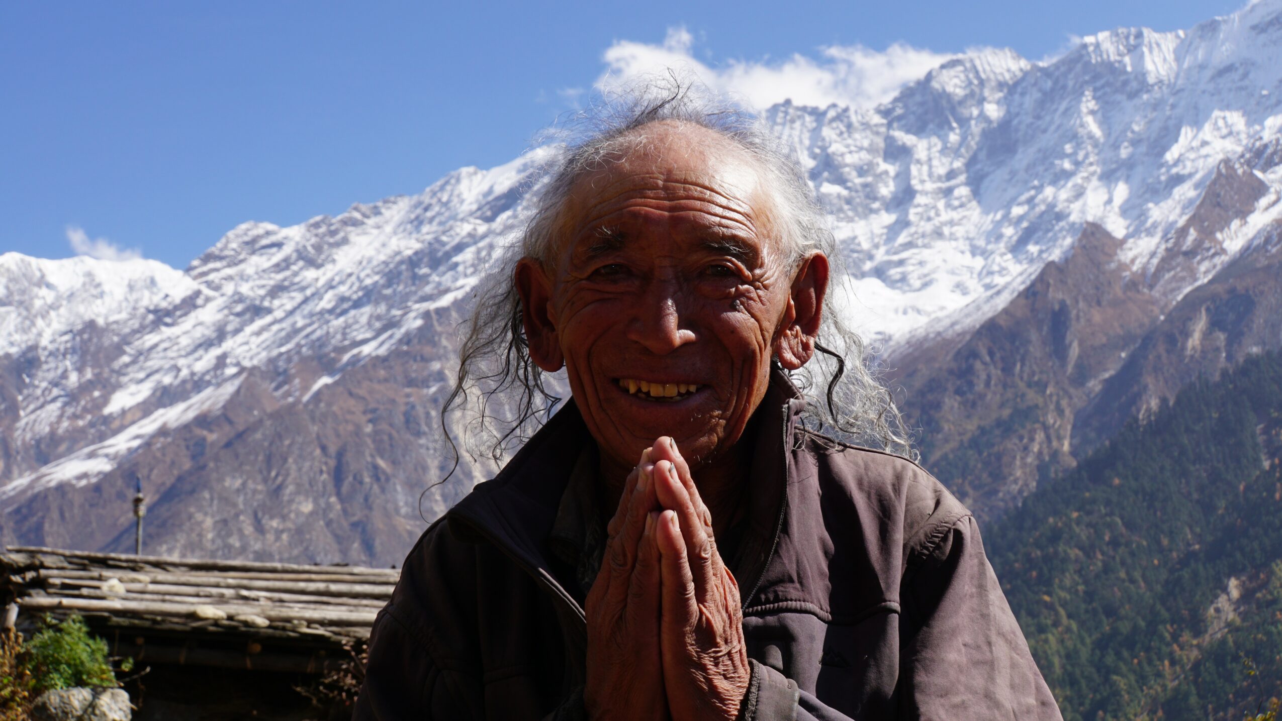 Elderly person with hands pressed together, smiling, with snow-capped mountains in the background.