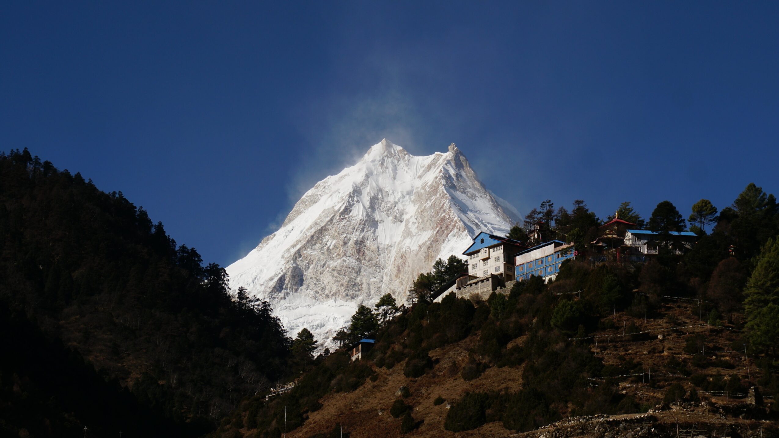 Snow-covered mountain with a clear blue sky, trees on hillside, and buildings on slope.