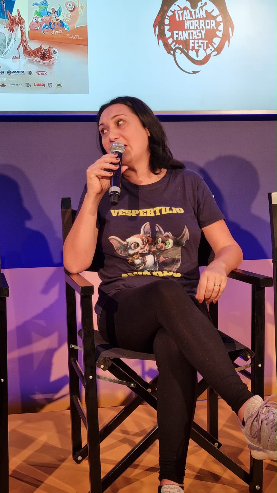Woman sitting on a chair, speaking into a microphone, wearing a dark T-shirt with a graphic, in front of a screen with a logo.
