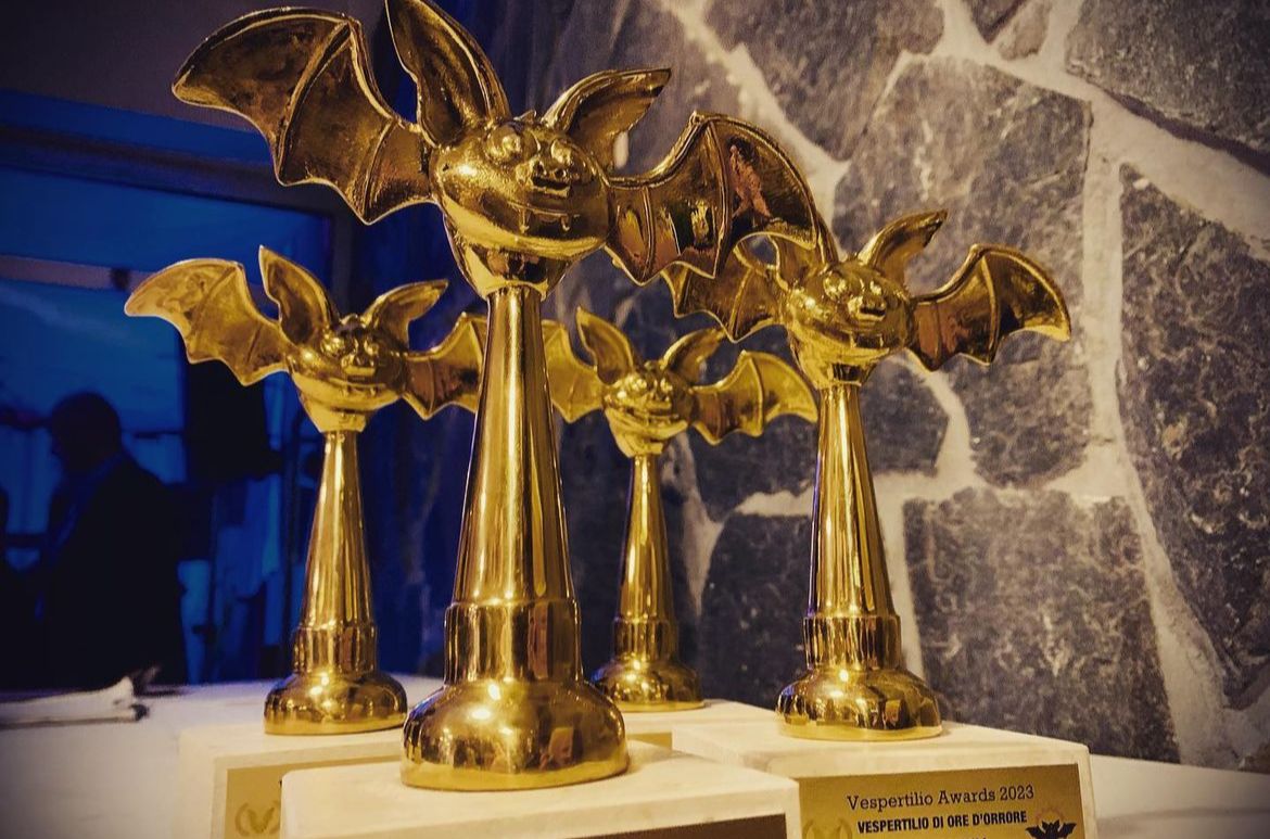 Four golden bat-shaped trophies on a white base against a stone wall background.