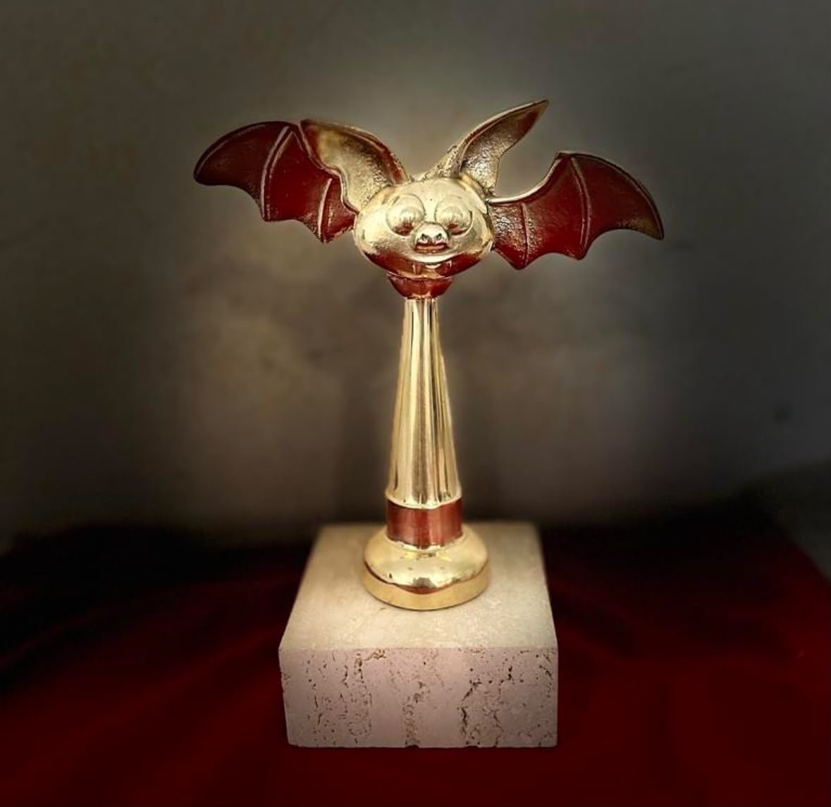 Decorative bat with a smiling face, wings, and a long neck, mounted on a white base, against a dark background.