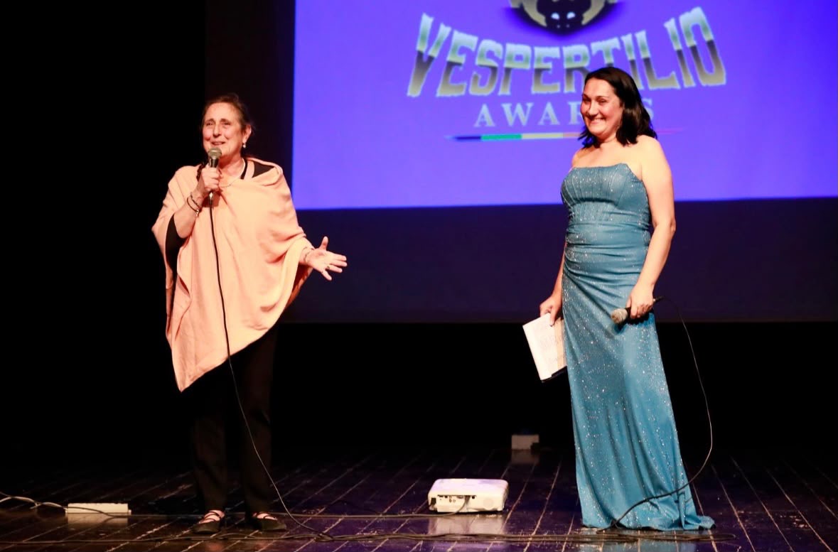 Two women stand on stage, one speaking into a microphone, the other holding a paper, with a large screen behind them displaying 'Vespertillo Awards'.