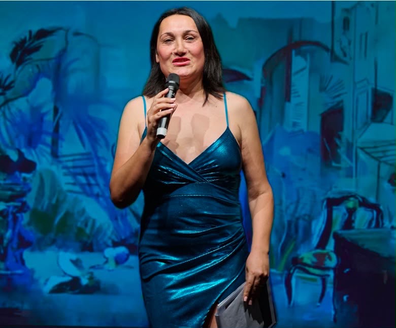 Woman in a blue dress holding a microphone on stage with a painted cityscape background.