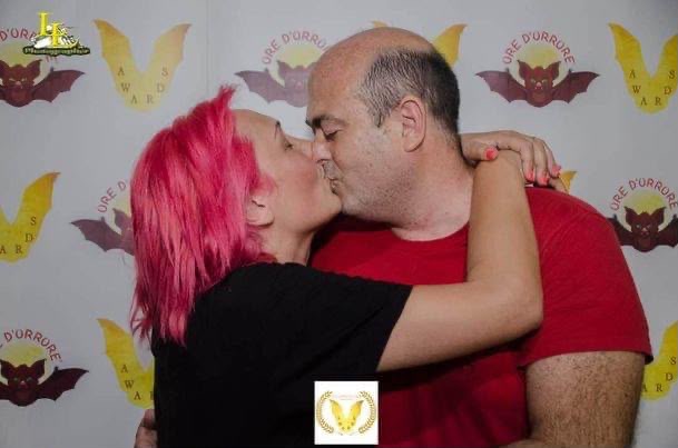 Two people kissing, one with pink hair and the other with a bald head, in front of a patterned background