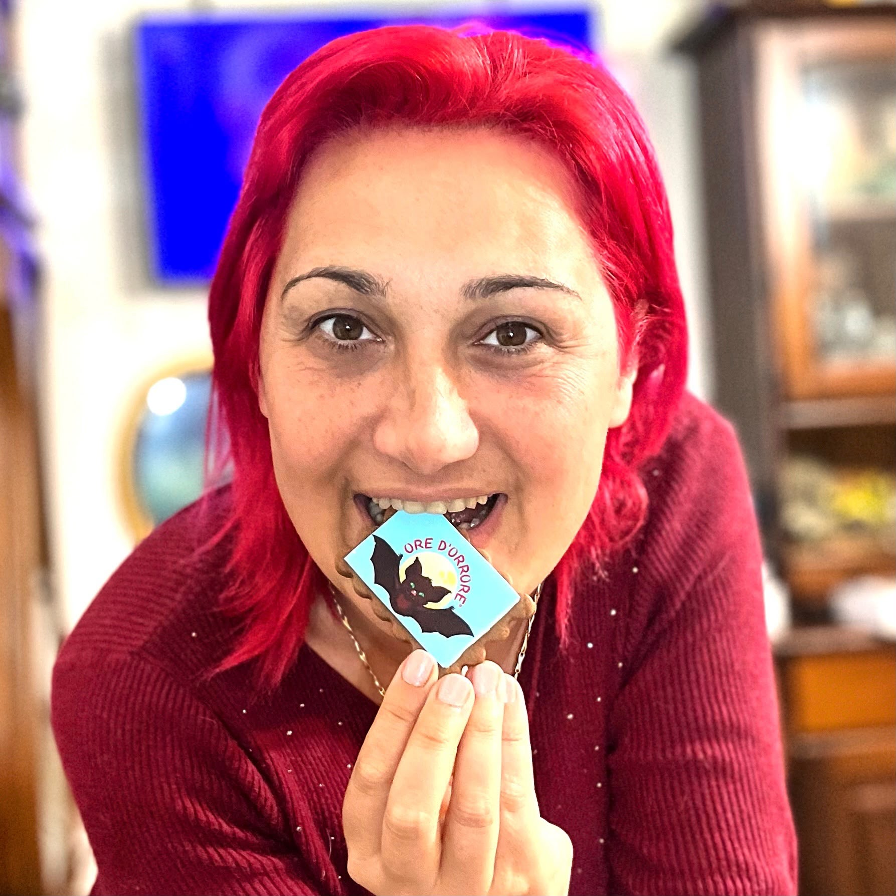 Woman with bright red hair smiling and holding a small card with a black cat illustration, indoors.