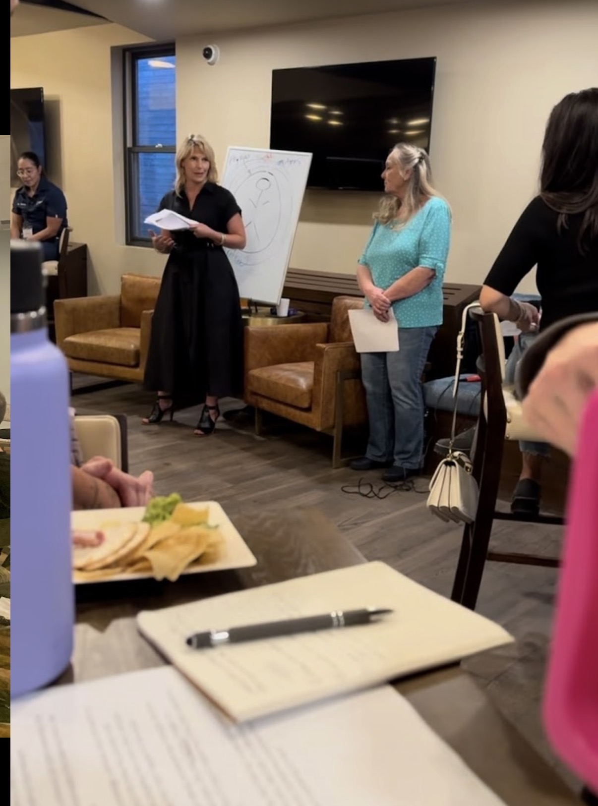 Three women standing in a room with a large TV, whiteboard, and chairs, engaged in a discussion or presentation.