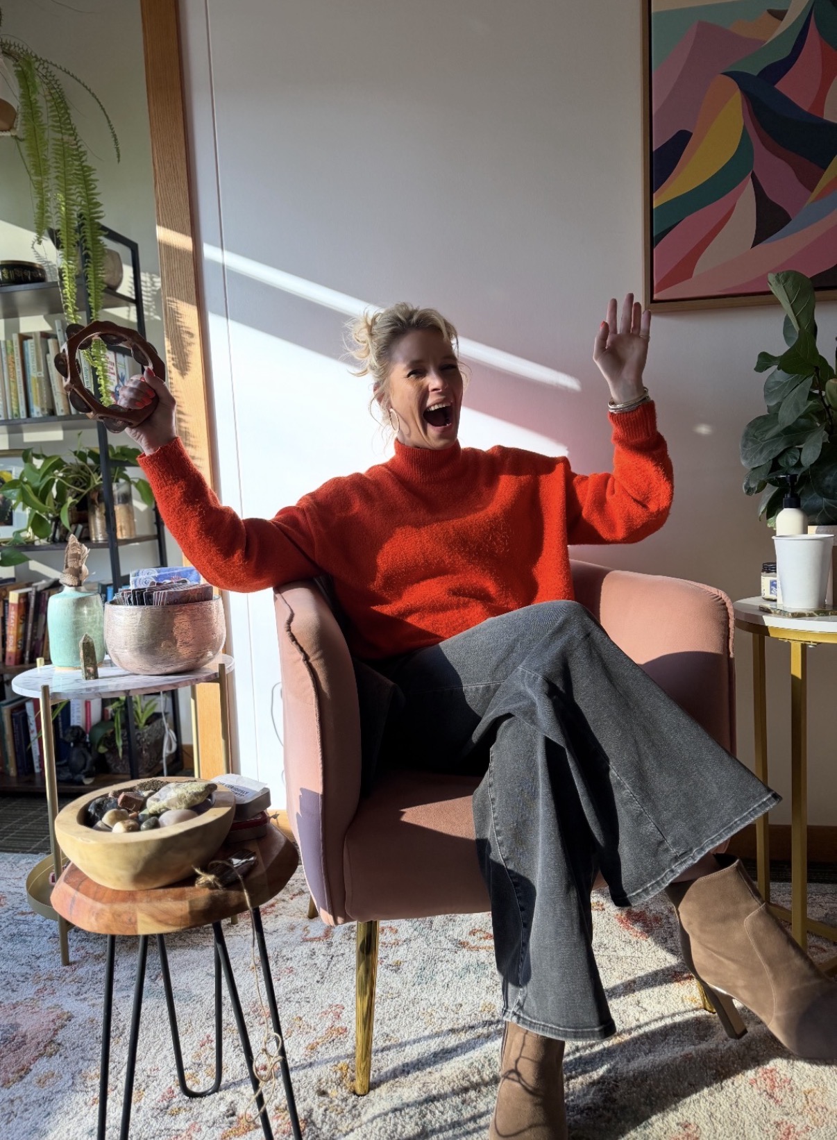 Woman sitting in a pink armchair, smiling and raising her right hand, wearing a red sweater and gray pants, in a room with plants and colorful artwork.