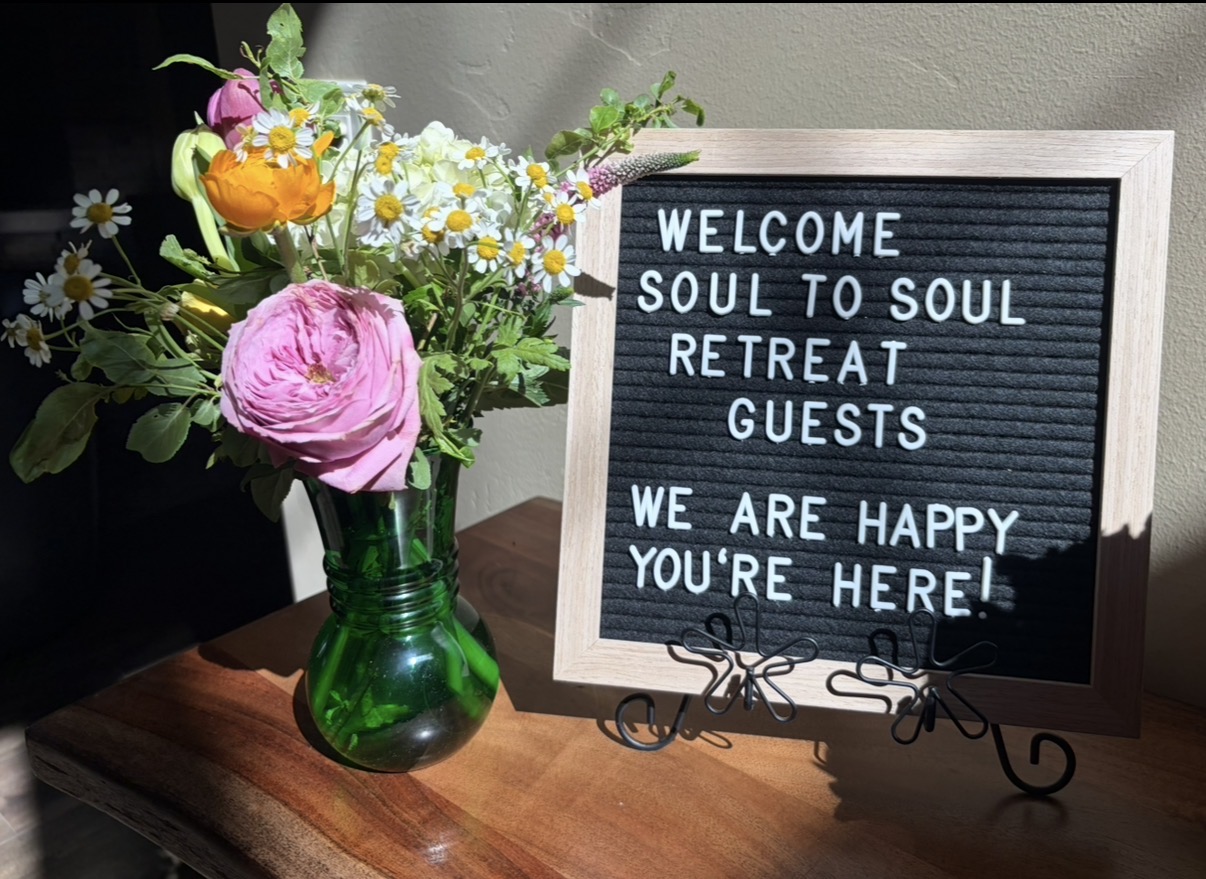 Vase with colorful flowers next to a welcome sign for a soul retreat, on a wooden surface.