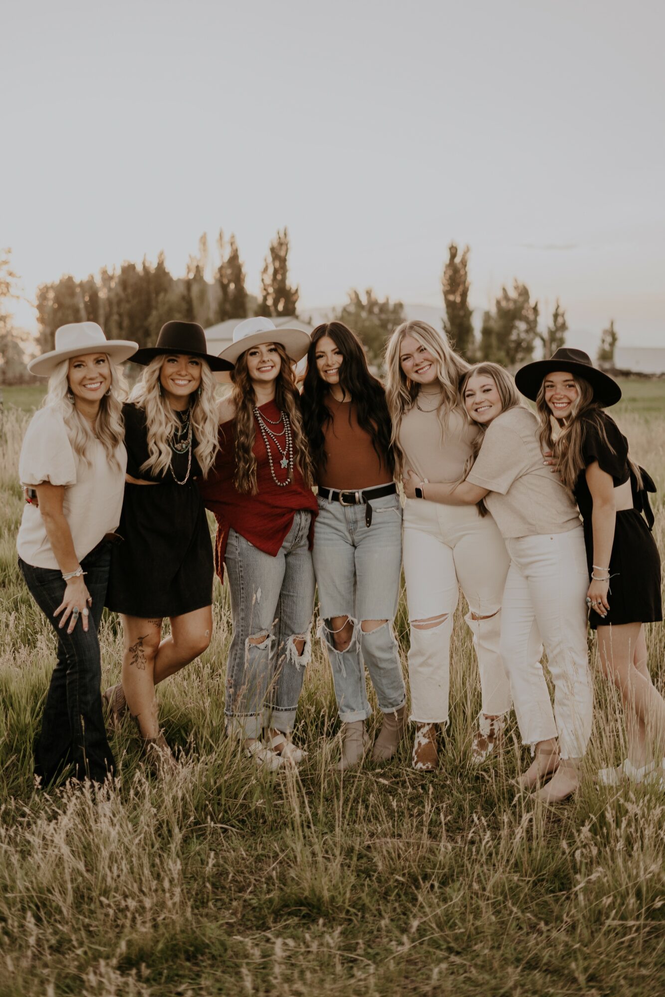 Group of seven women standing outdoors in a field, smiling, wearing casual clothing and hats, with trees in the background.