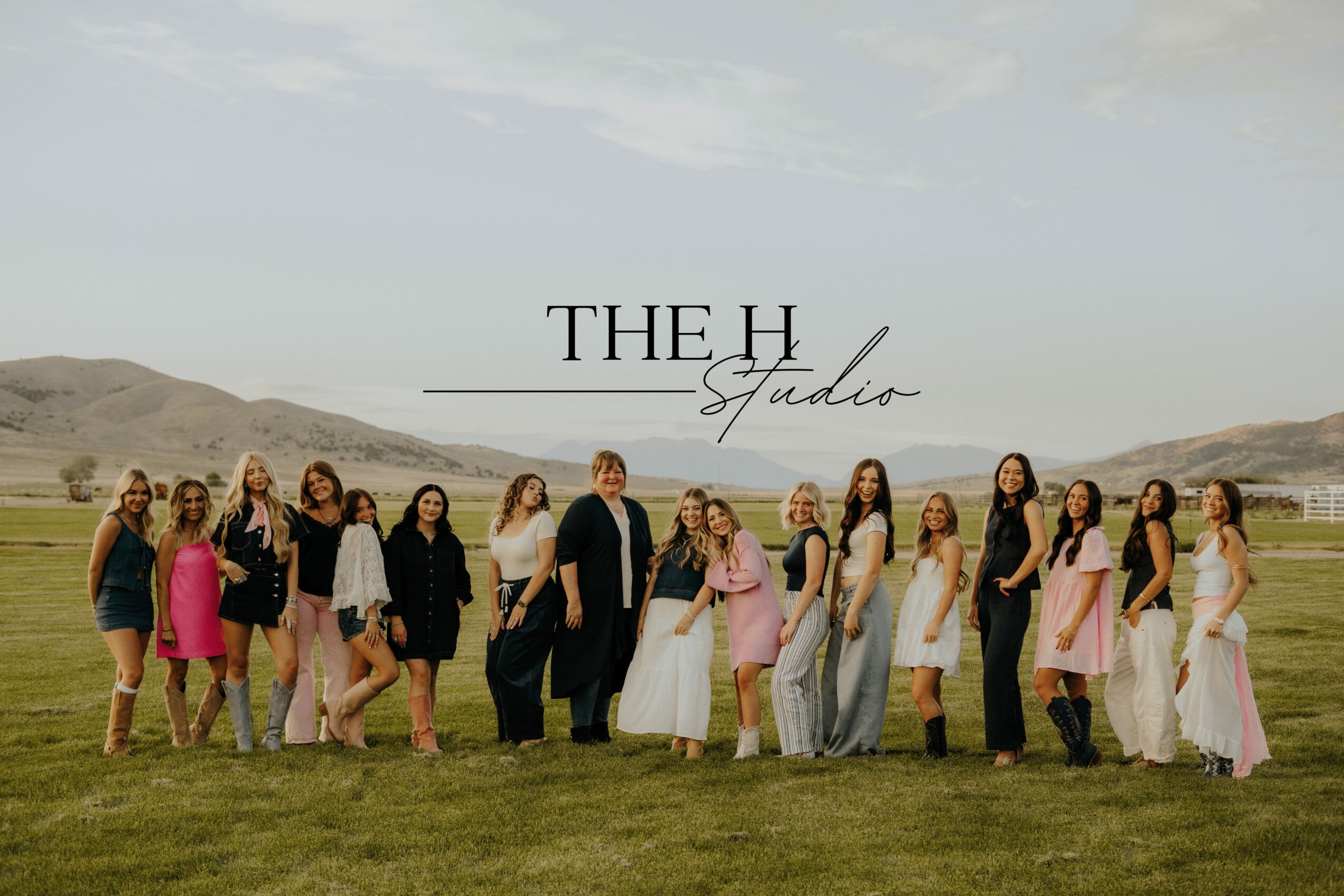 Group of people standing outdoors on grass with hills in background, some in formal attire, posing for photo.