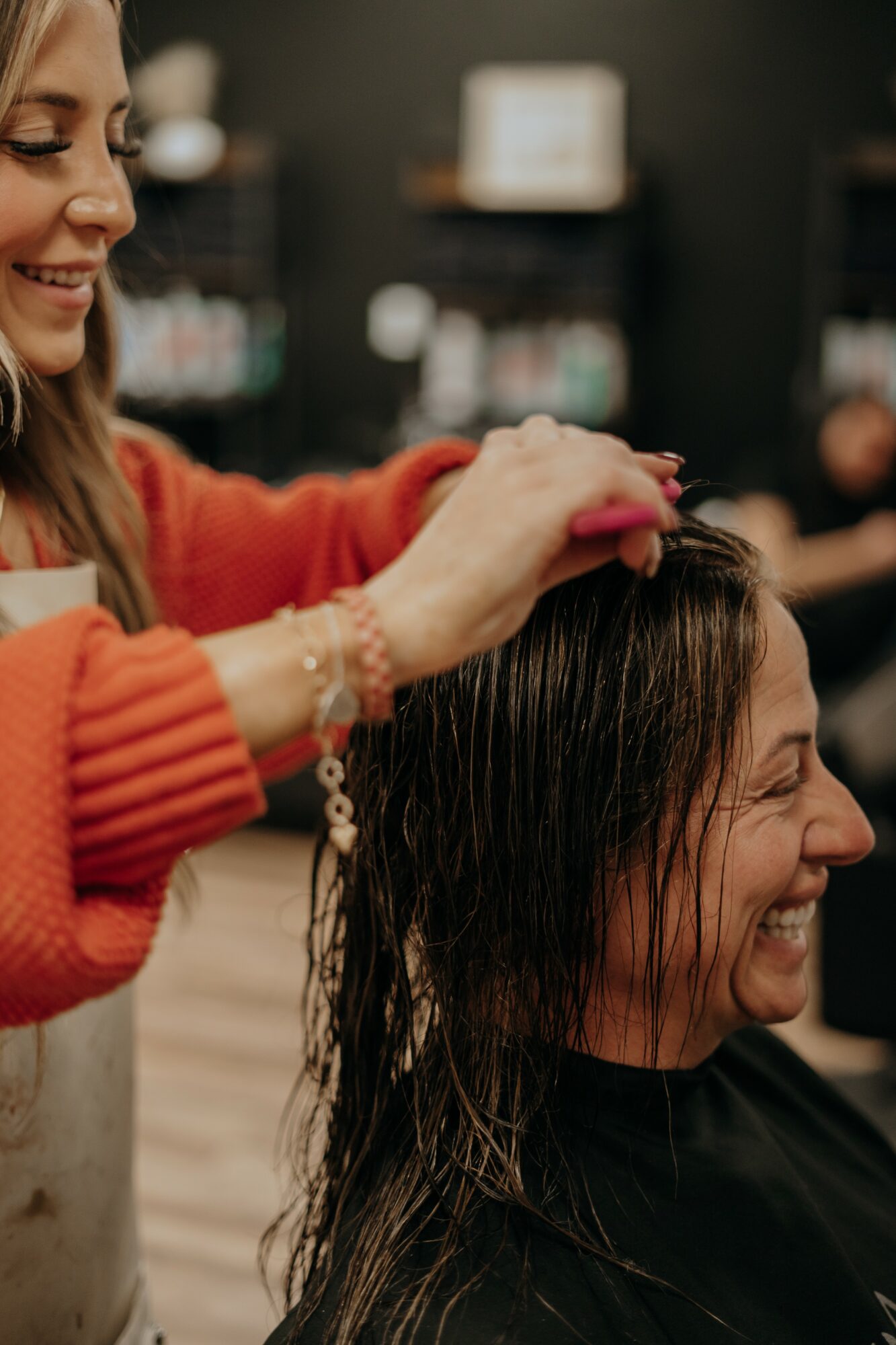 Hairdresser styles a woman's wet hair in a salon, smiling and wearing an orange sweater.
