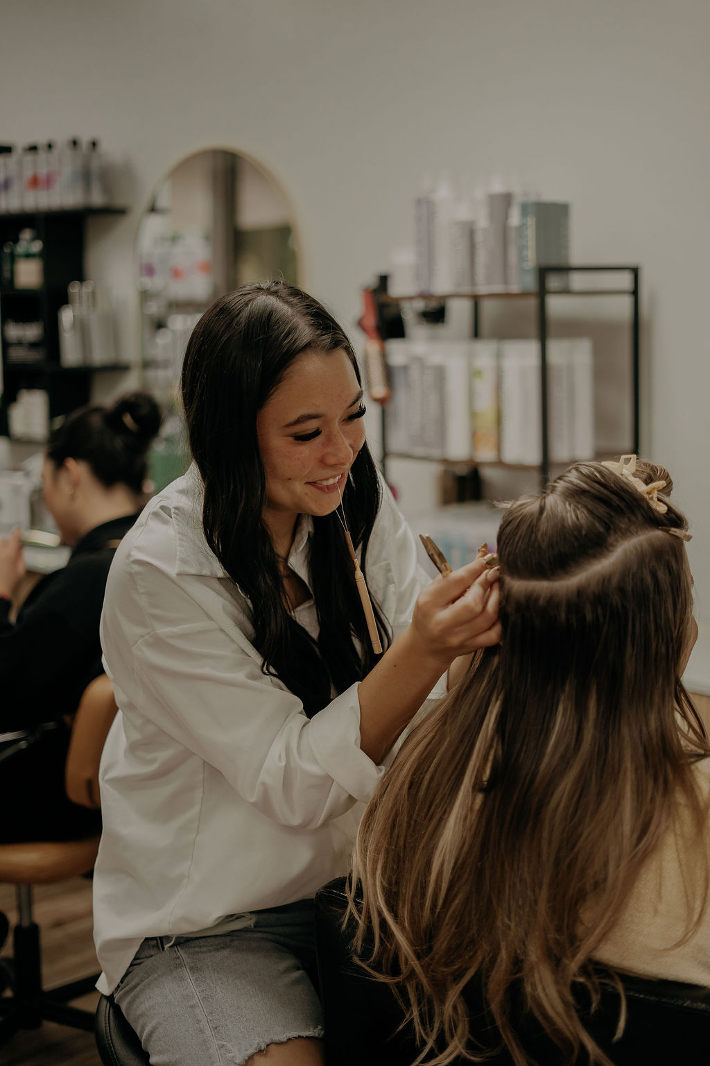 A woman with long dark hair styles a person's hair in a salon, smiling and focused on her work.