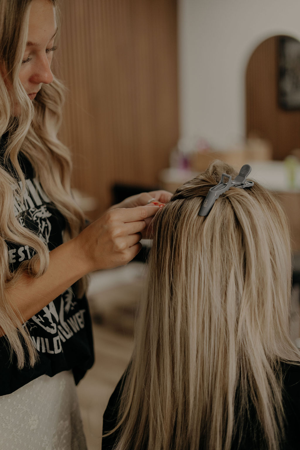 Woman styling a person's blonde hair with a bow, in a room with wooden and white walls.