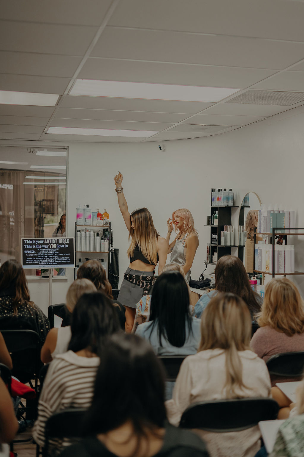 Two women stand in front of seated audience in a room with shelves and products, one raising her hand.