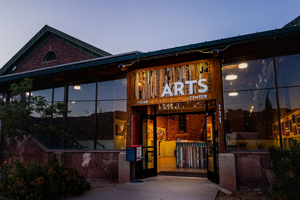 Arts center entrance with glass windows and a sign reading 'ARTS' above the door, brick building reflections, evening lighting.