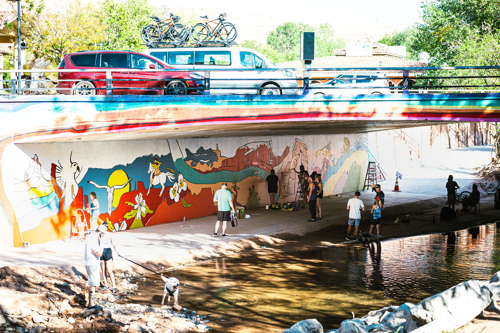 People painting a colorful mural under a bridge, with cars parked above and trees in the background.