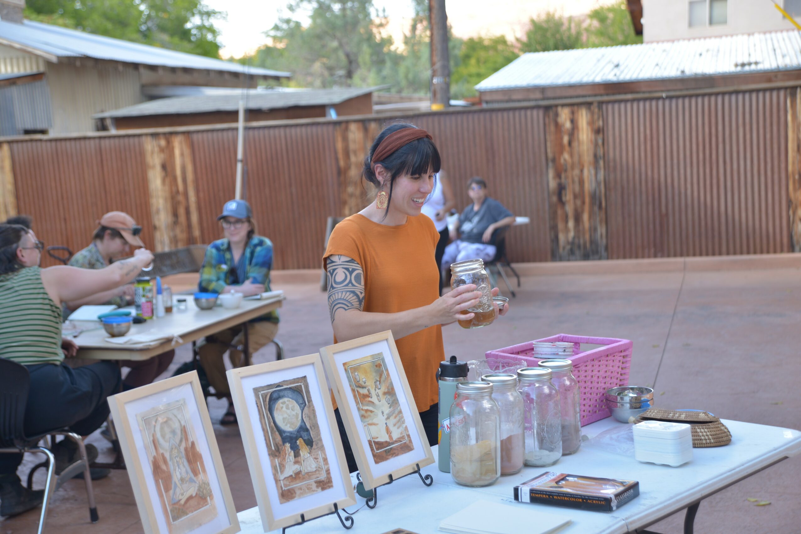 Young woman with dark hair and tattoos on her arm stands at a table outdoors, holding a glass jar, with people sitting at a table in the background.