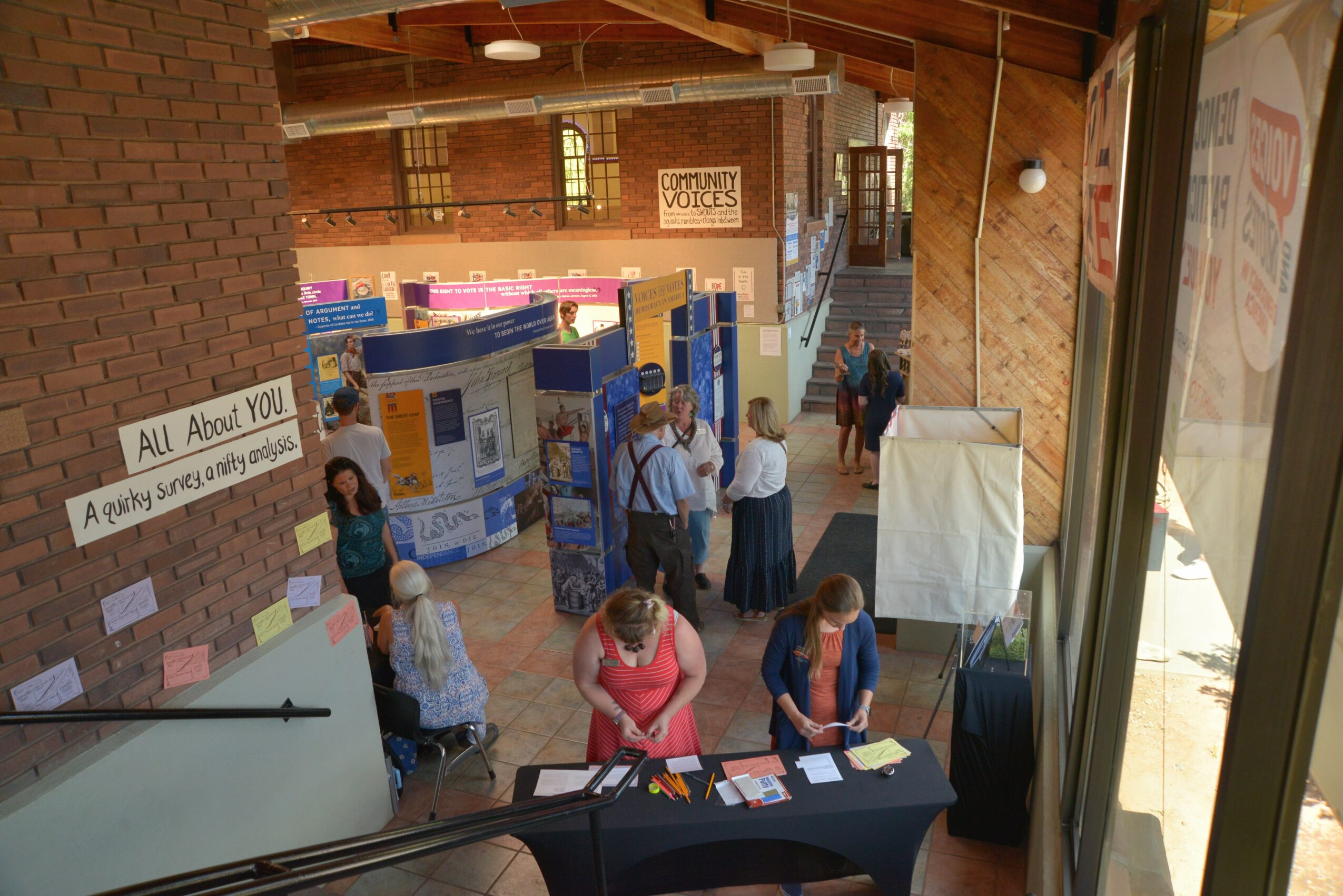 People at an indoor exhibition with display boards and informational booths, viewed from a high angle near a window.