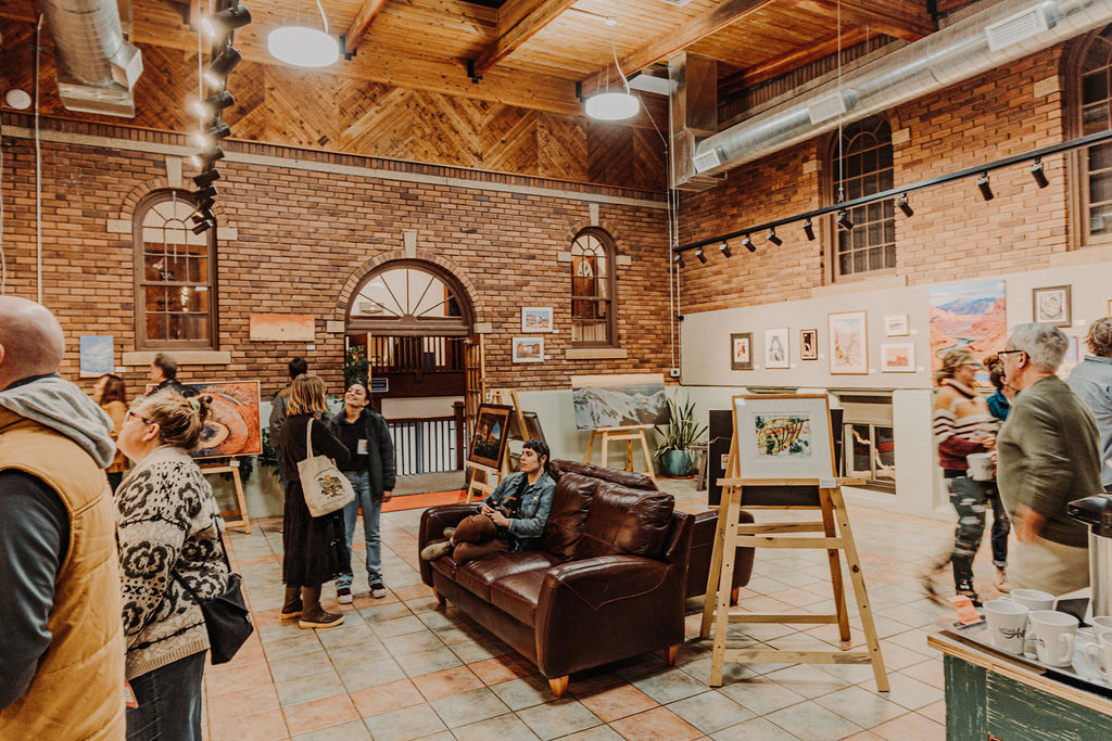 People gather in an art gallery with brick walls, wooden ceiling, and framed artwork displayed on walls and easels.