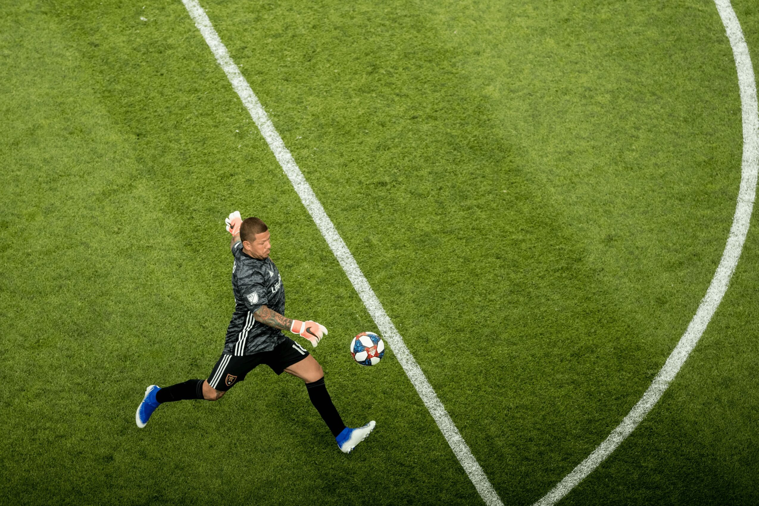 Soccer goalkeeper in black uniform running on field with ball, viewed from above.