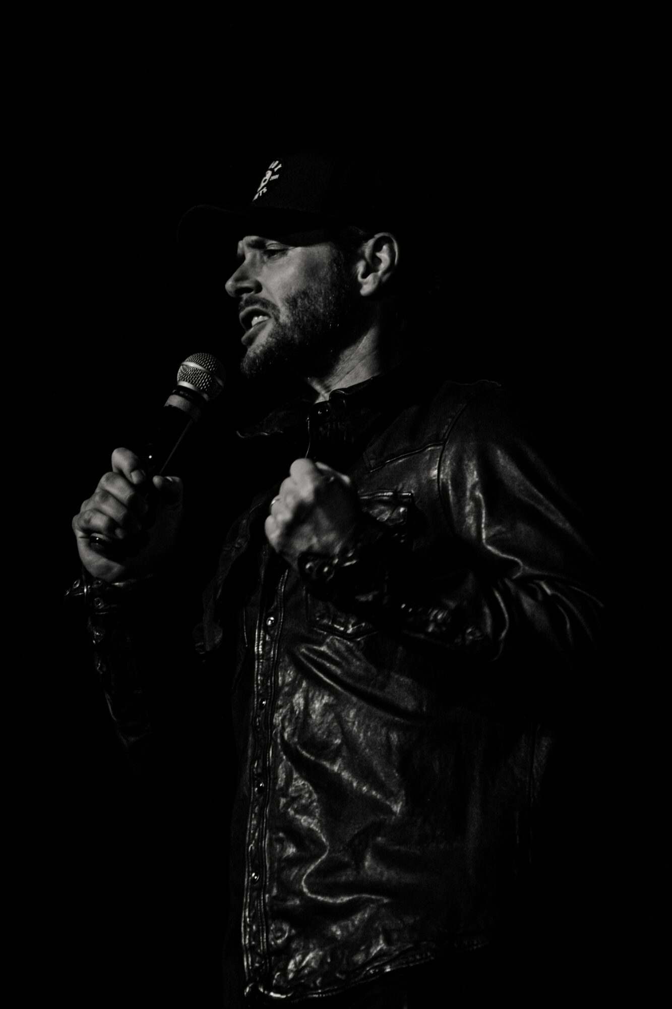 Man wearing a cap and leather jacket holding a microphone, speaking or performing on stage, in black and white.
