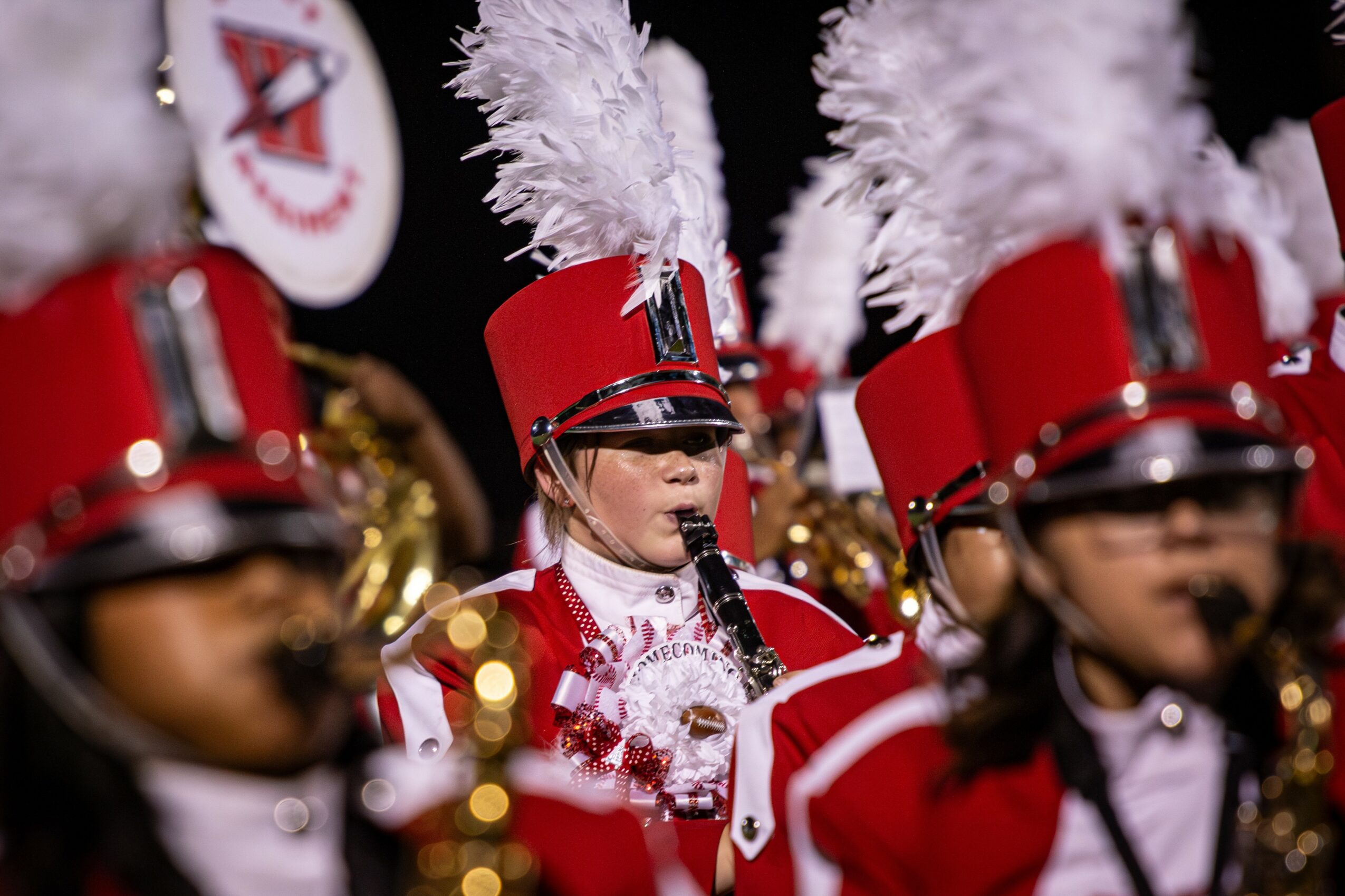 Children in marching band uniforms with red hats and white plumes perform outdoors.