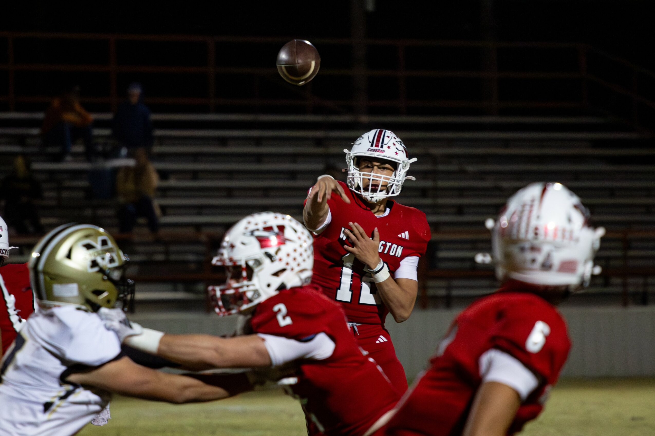 Football player in red jersey throws ball, teammates and opponent in white jersey reach for it on field, stadium background.