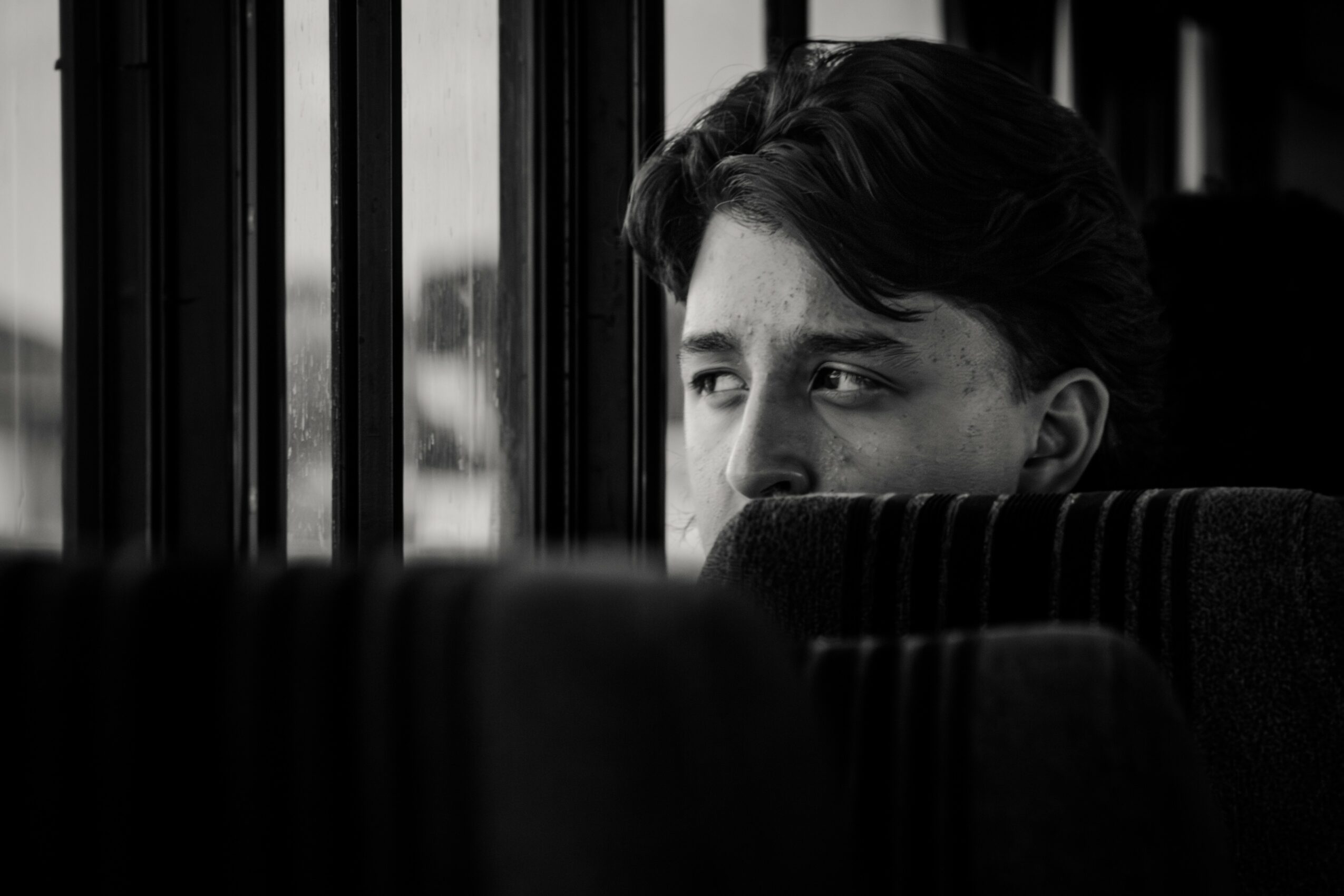 Young person looking out of a bus window, partially obscured by seat backs, in black and white.