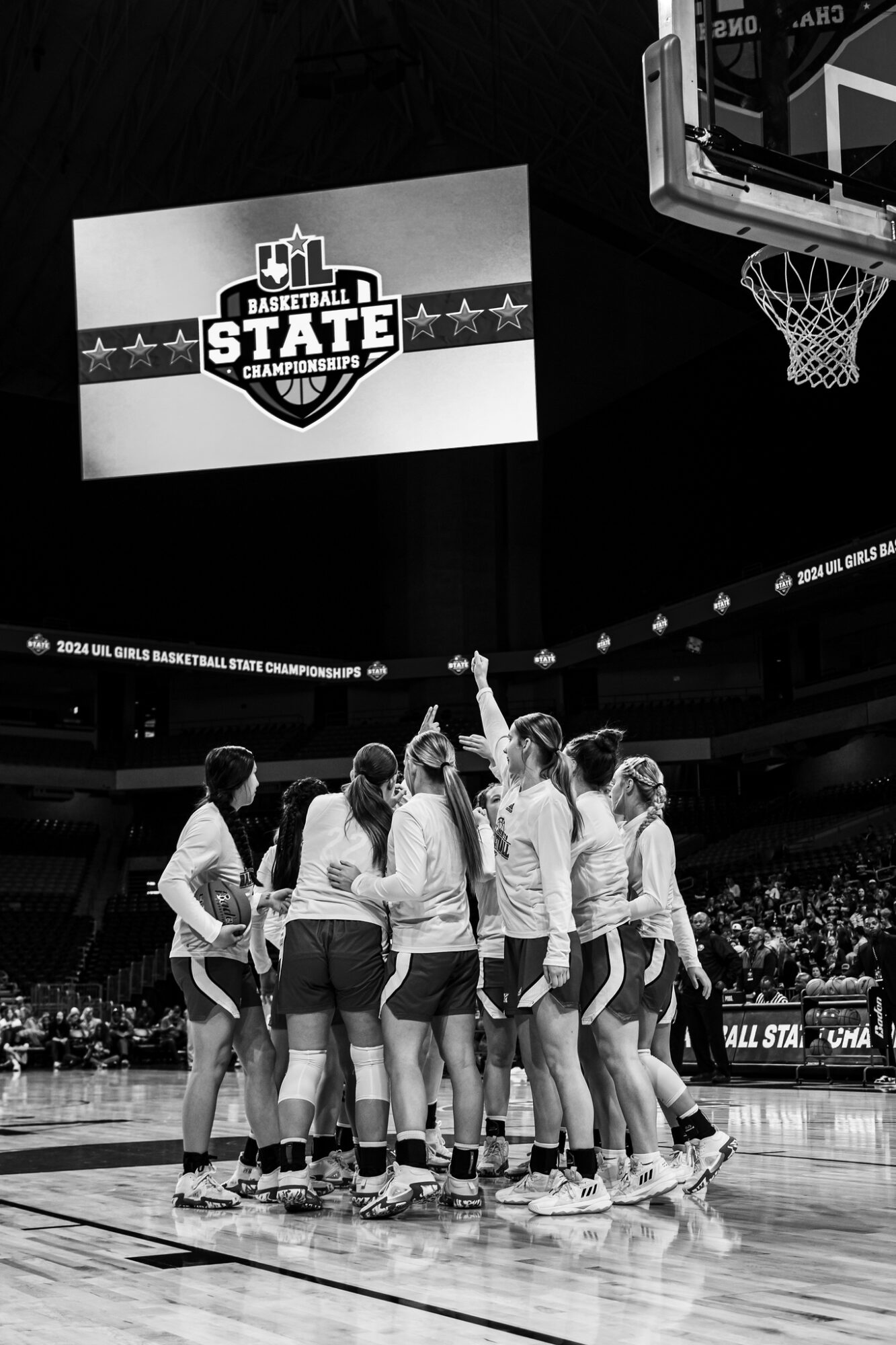 Group of basketball players huddled on court, with a scoreboard and basketball hoop visible in the background.