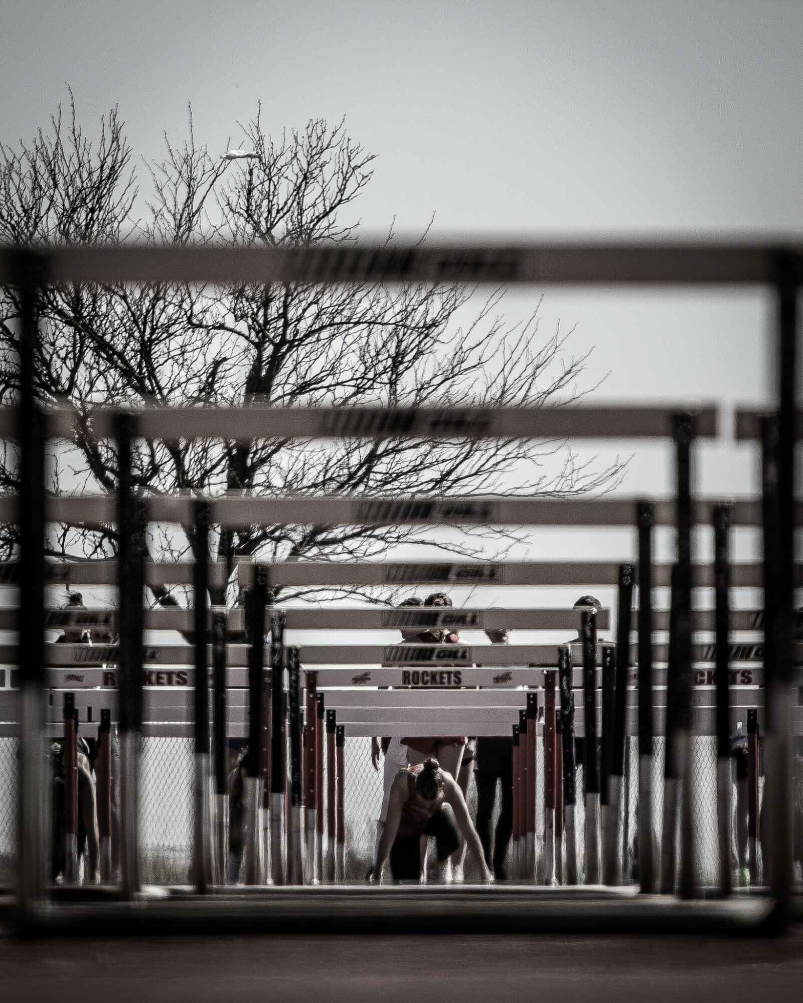 Person bending over under a series of horizontal bars outdoors, with leafless trees in the background.