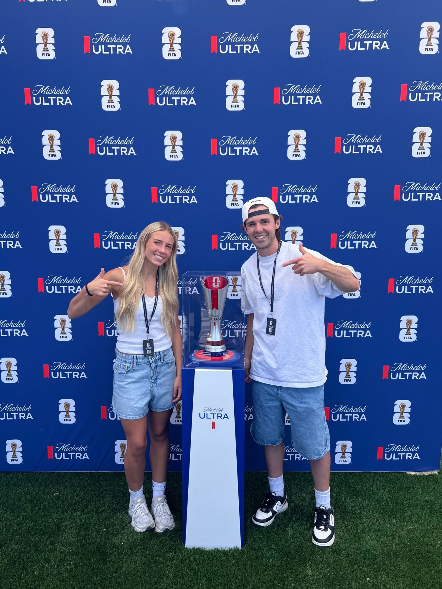Two people standing on a podium with a blue backdrop, one holding a trophy, the other making a peace sign, outdoors.