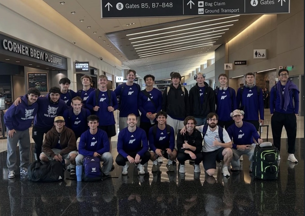Group of young people in blue jackets posing in an airport terminal, some kneeling and some standing, with luggage.