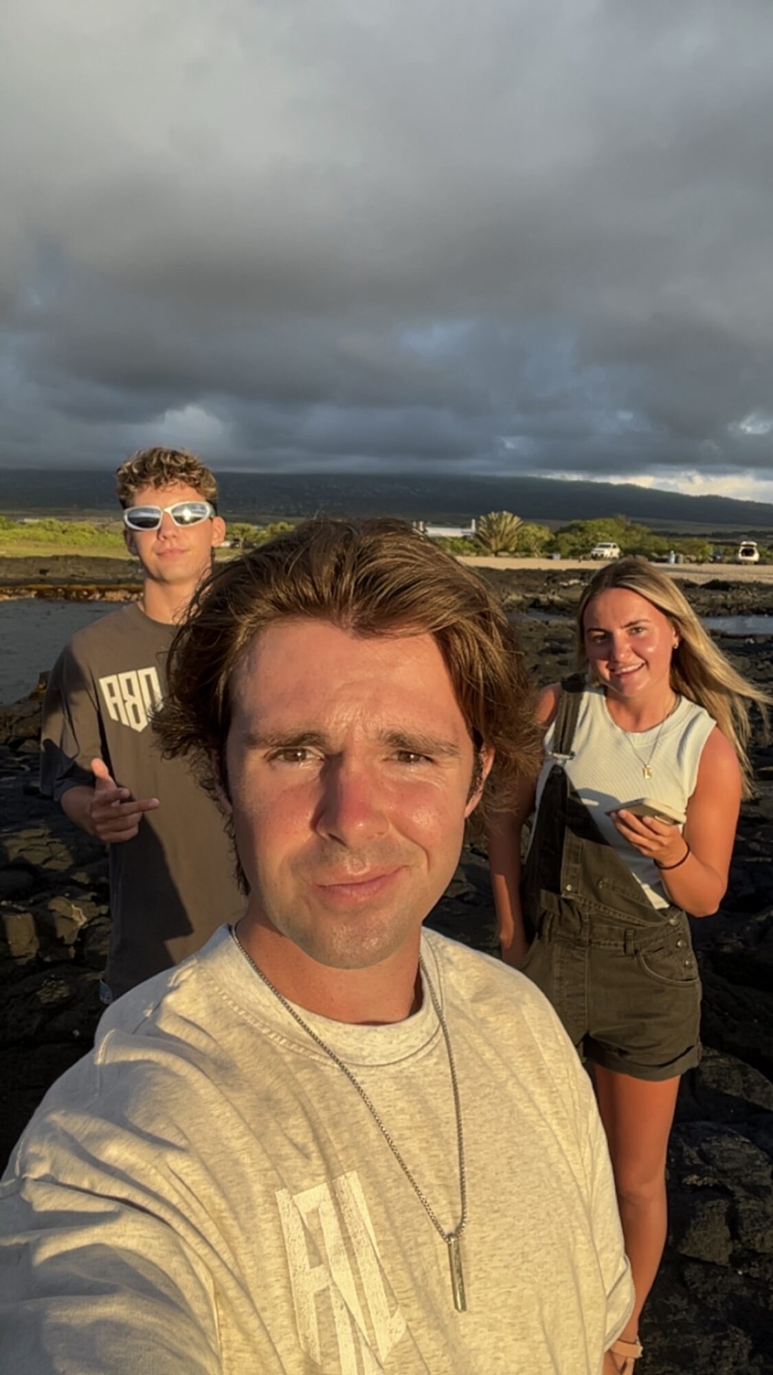Three people outdoors with cloudy sky, standing on a stone surface, smiling at the camera.