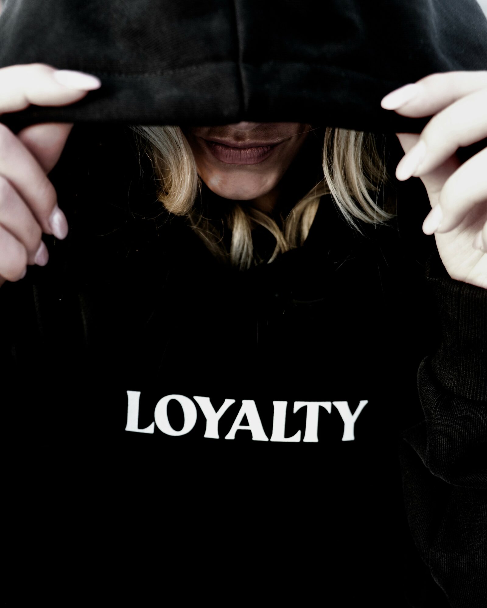 Person wearing a black hoodie with the word 'LOYALTY' on it, pulling the hood over their face, smiling slightly.